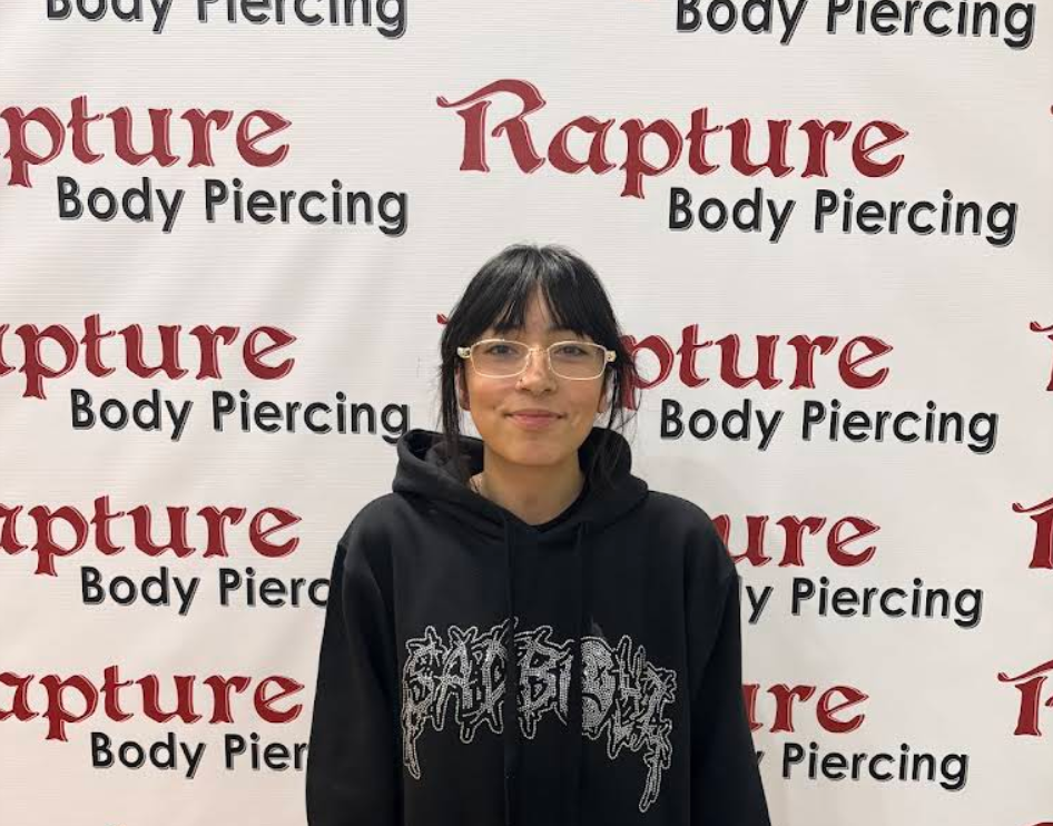 A woman with glasses and black hair standing in front of a backdrop with the words 'Rapture Body Piercing' repeated on it.