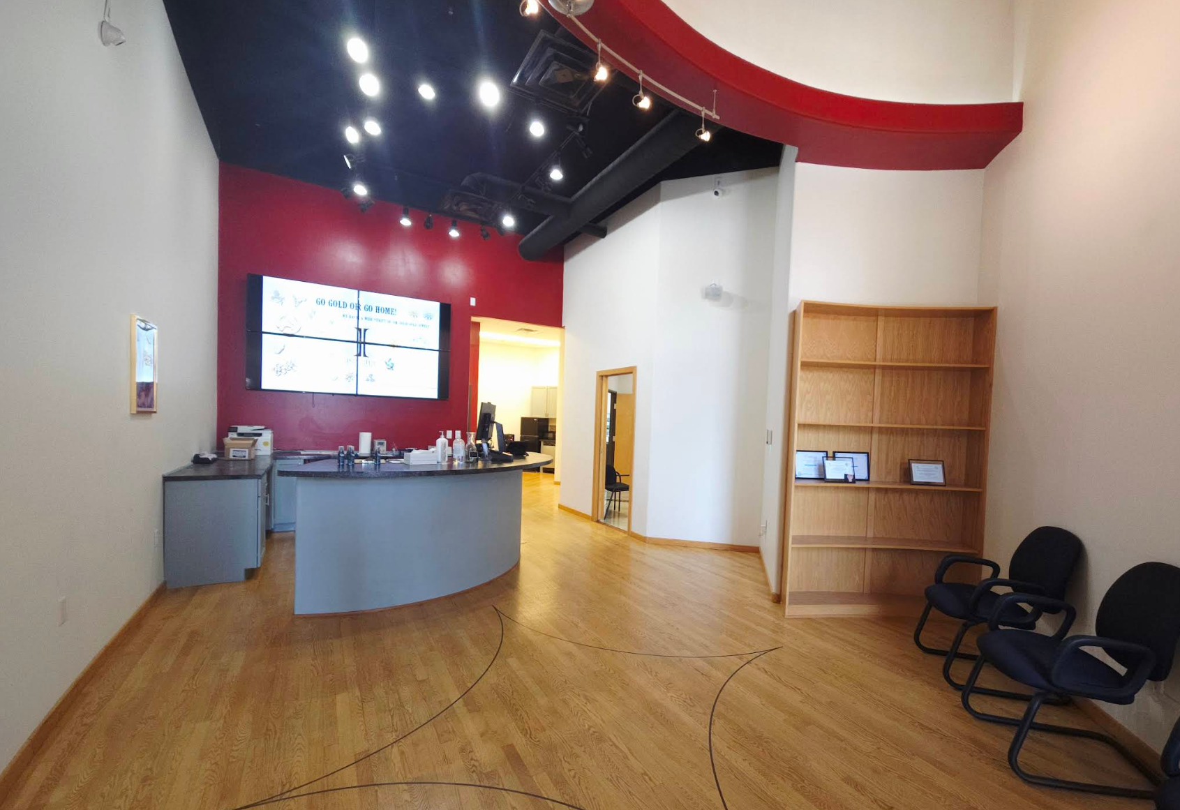 Waiting area with black chairs, wooden shelves with framed documents, a reception desk with bottles and supplies, a large LED display on a red wall, and a doorway leading to an office area.