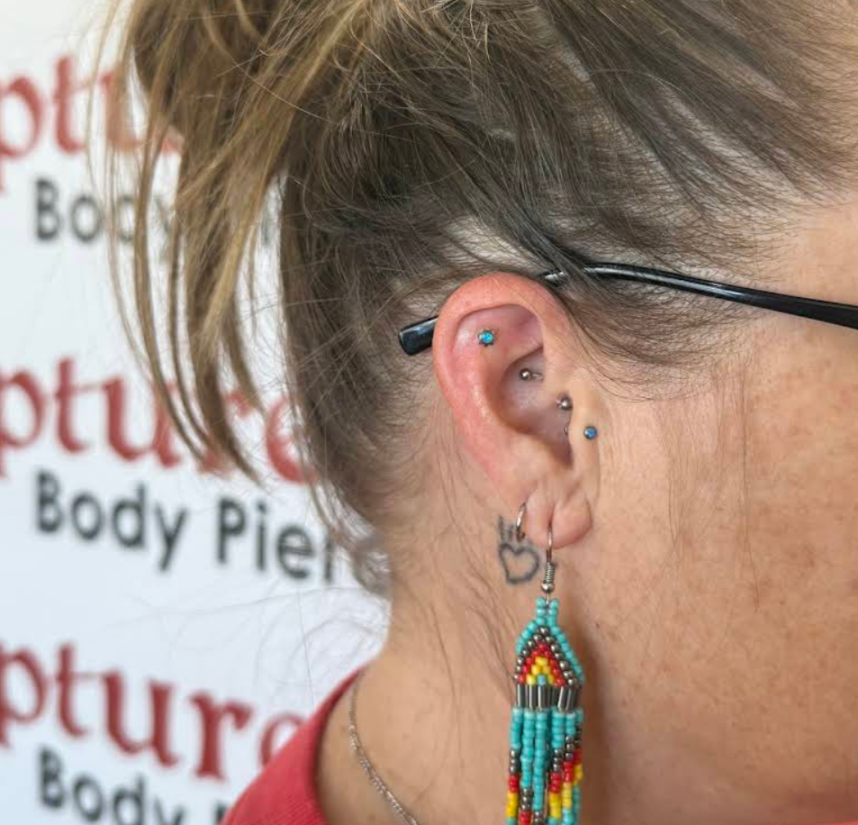 Close-up of a woman with multiple earrings, including a blue gem piercing on her upper ear, a small silver hoop, and a colorful beaded earring with a rainbow pattern. She is wearing glasses and has light brown hair tied back, with a partial view of a