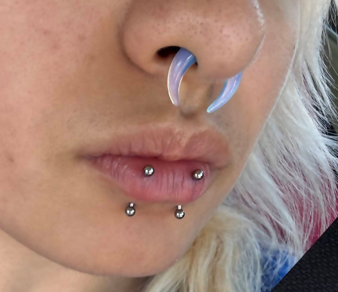 Close-up of a person's face showing their nose, lips, and multiple facial piercings, including a septum piercing with a clear purple septum ring and two lip piercings with silver balls.