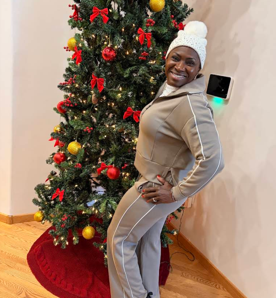 A smiling woman in a beige tracksuit and a white knit hat stands next to a decorated Christmas tree.