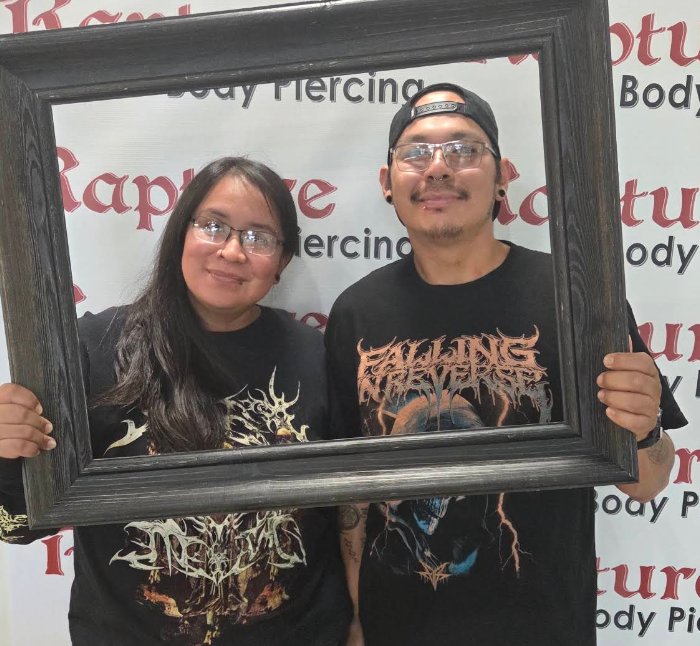 A man and a woman are holding a black picture frame, posing for a photo at a tattoo and body piercing studio. The woman has long dark hair and glasses, and the man wears glasses and a backward baseball cap. Both are wearing black graphic t-shirts.
