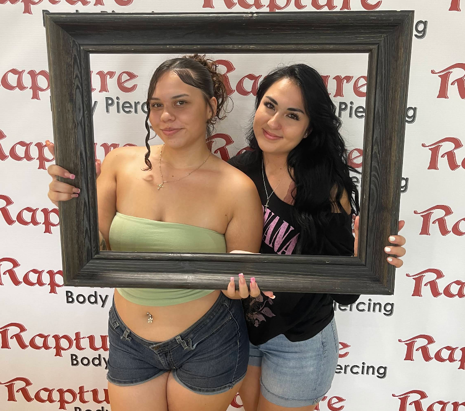 Two women posing with a large wooden picture frame, standing in front of a backdrop with the words 'Rapture Body Piercing' repeatedly printed in red and black.