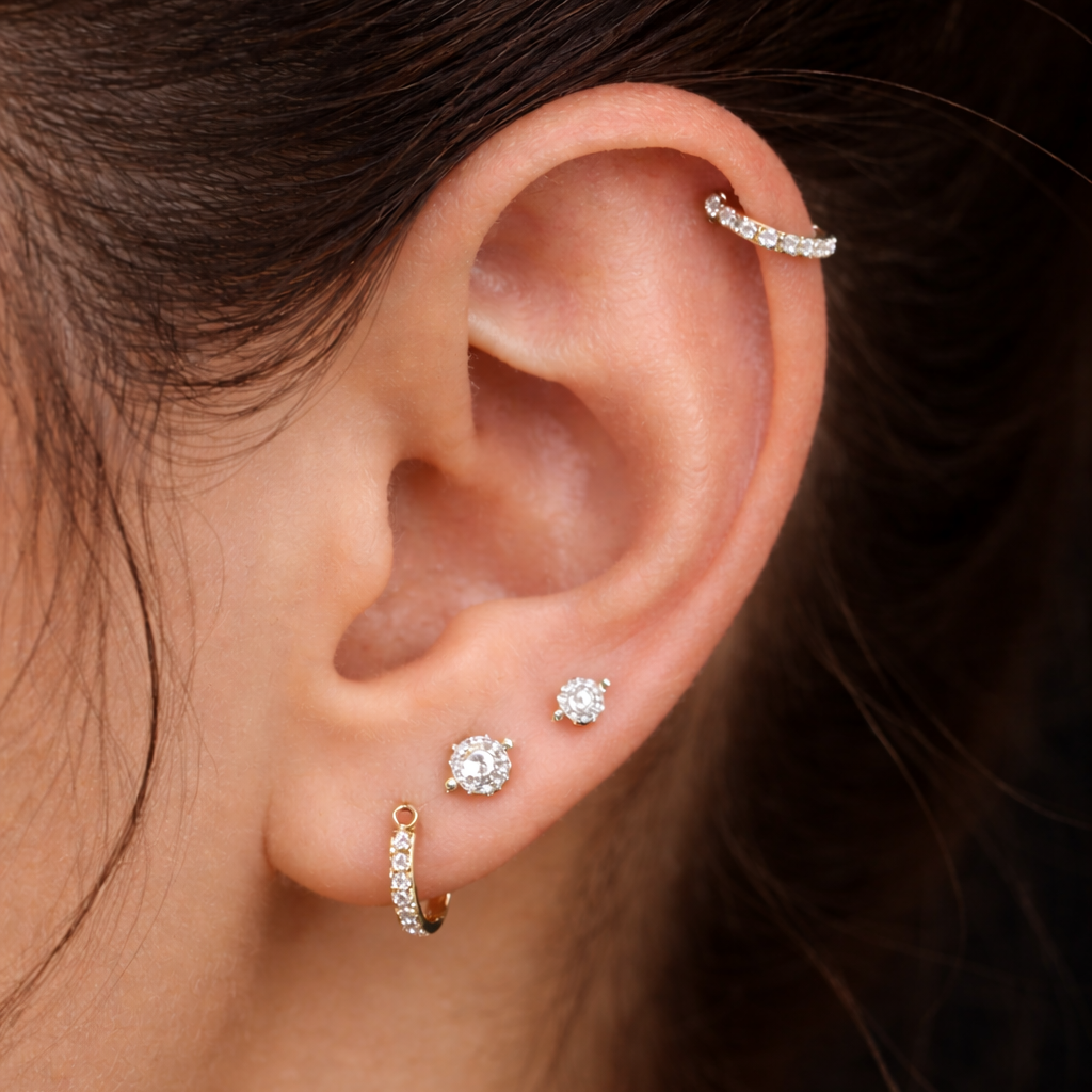 Close-up of a woman's ear with multiple earrings, including a gold hoop and three studs with clear stones.