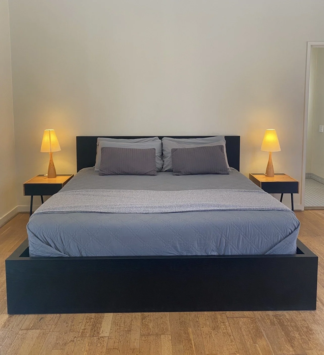 A modern bedroom with a large bed, grey bedding, two pillows, two bedside tables with yellow lamps, and a visible doorway to a bathroom in the background.