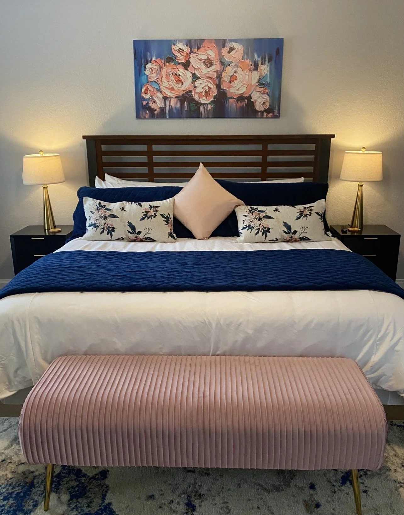 A neatly made bed with navy blue, white, and blush pink pillows, a navy quilt, bedside lamps on black nightstands, and a floral painting above the headboard. A pink velvet ottoman with gold legs is in front of the bed.