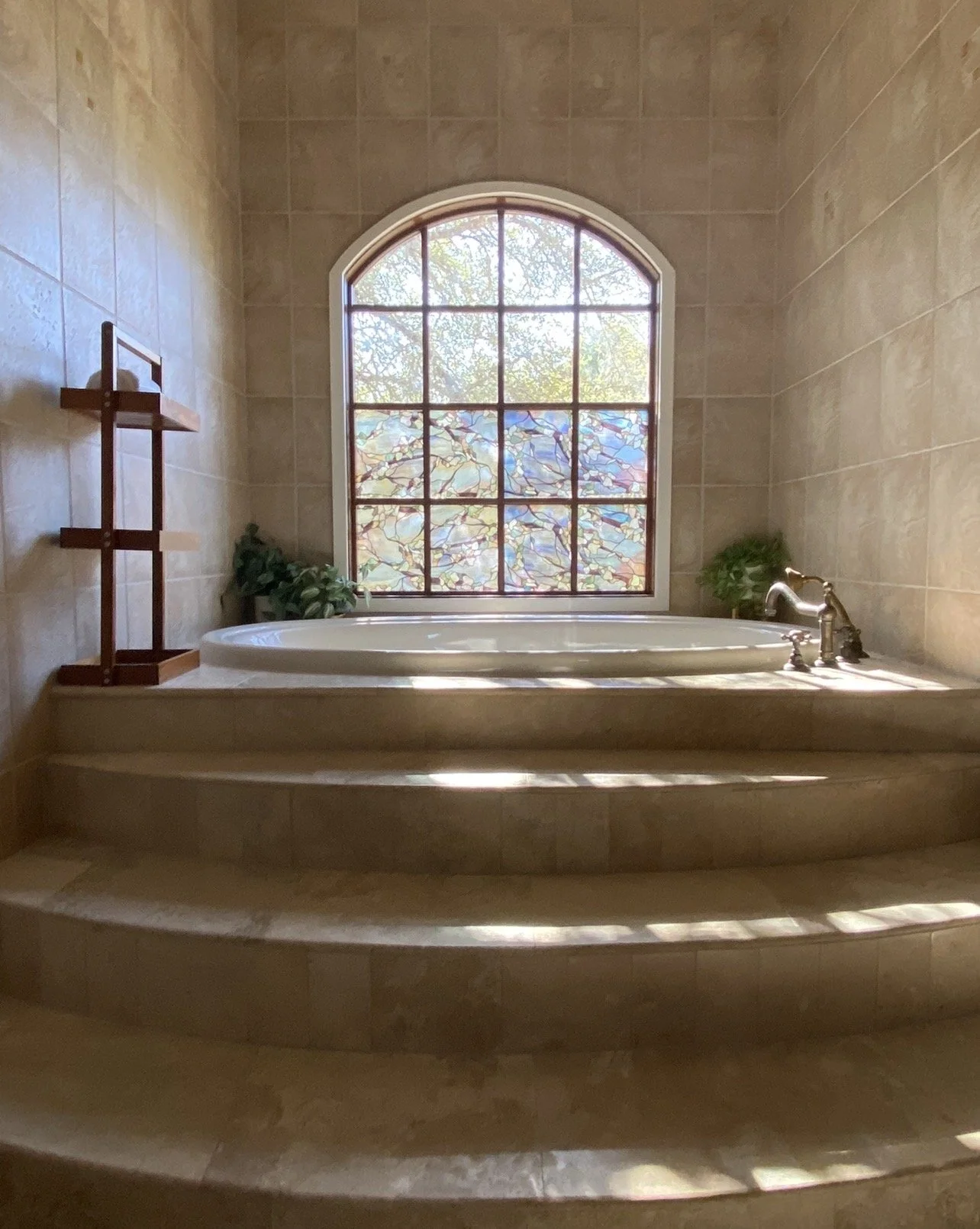 A bathtub in a bathroom with beige tiled walls and steps leading up to it. There is a large arched window with stained glass, plants on each side of the bathtub, and a wooden shelf on the left wall.