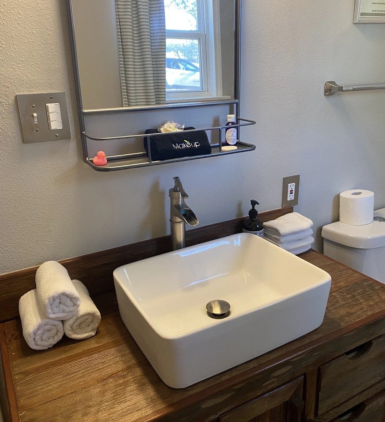 A bathroom vanity with a white vessel sink, a modern faucet, and rolled white towels on a wooden countertop. Above is a mirror with a black makeup bag, hand soap, and small decorative items. To the right is a toilet with a paper towel roll on top. The wall has a window with a striped curtain, and an electrical outlet with a plug and a USB port is visible.