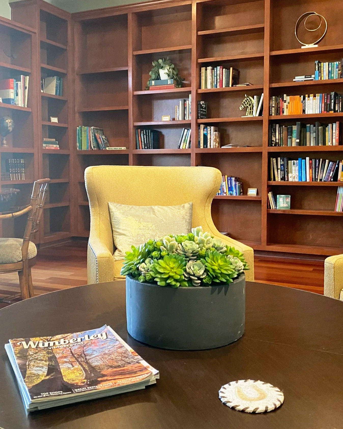 A cozy library or reading room with a yellow armchair, a bookshelf filled with books, a dark wooden table with a succulent plant in a gray pot, a magazine titled 'Wumberley,' and a small decorative paper plate. The background features a large wooden bookshelf with multiple shelves and decorative items.