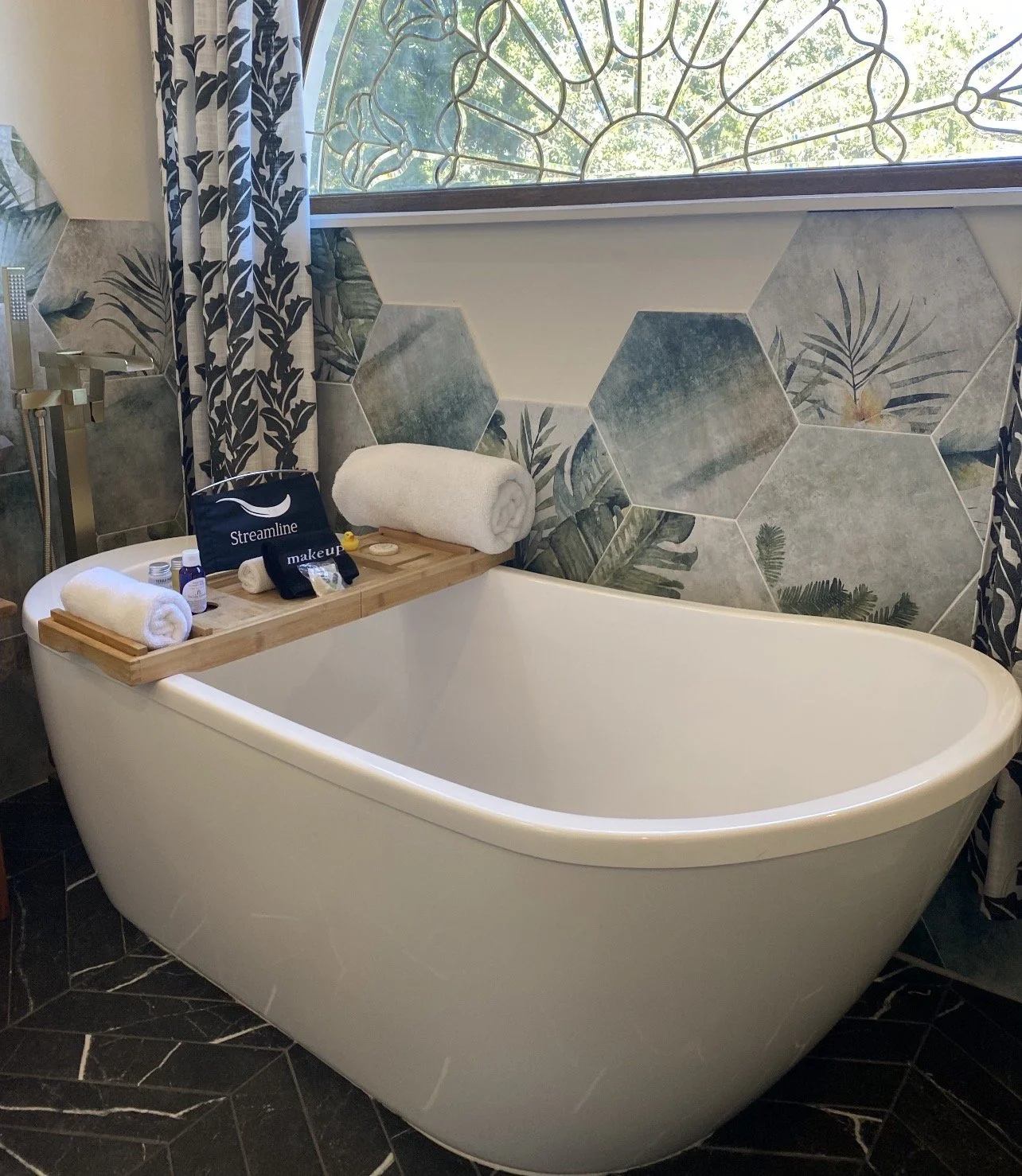 Bathroom with a white oval bathtub, a wooden tray holding rolled towels and toiletries, and a decorative wall with hexagonal tiles featuring tropical leaf patterns. There is a window with ornate ironwork and curtains with a leafy design.