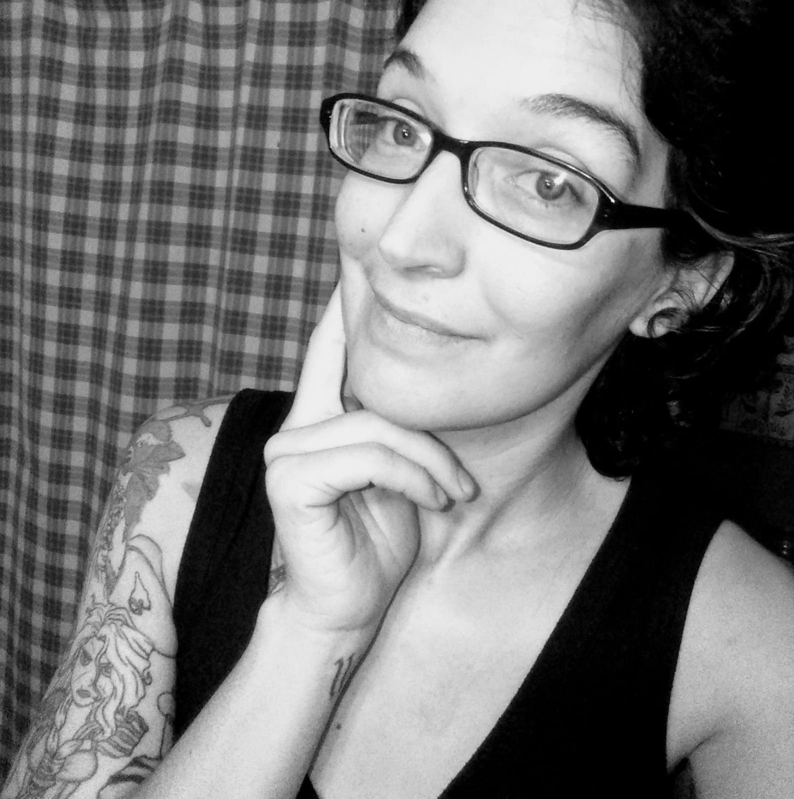 A woman with short dark hair and glasses, smiling, with her chin resting on her hand. She is wearing a sleeveless black top and has a tattoo on her left arm. The background features a checkered curtain.