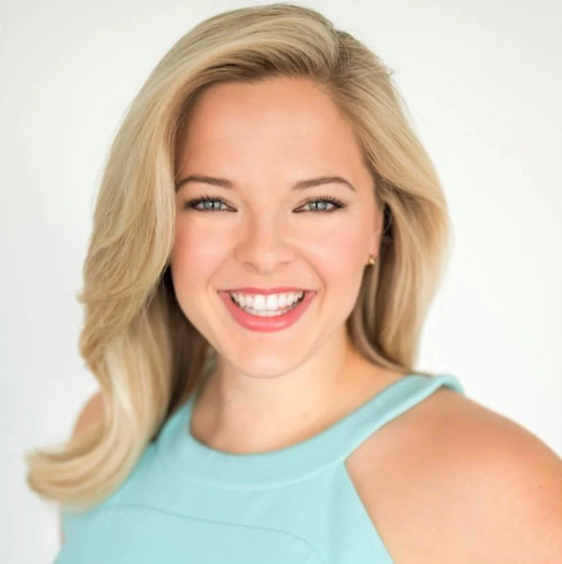 A woman with blonde hair smiling, wearing a light blue sleeveless top against a plain white background.