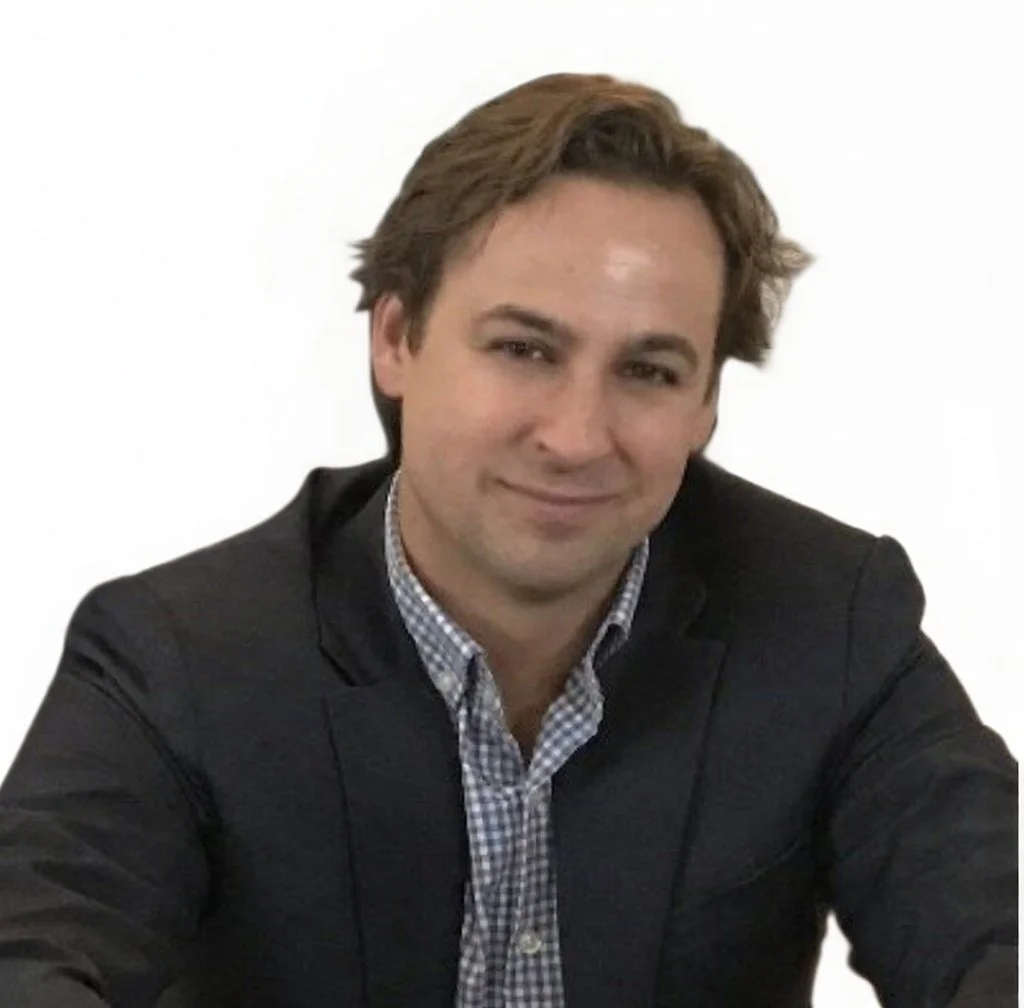 A man with brown hair styled back, wearing a dark blazer and checked shirt, smiling at the camera, against a plain white background.
