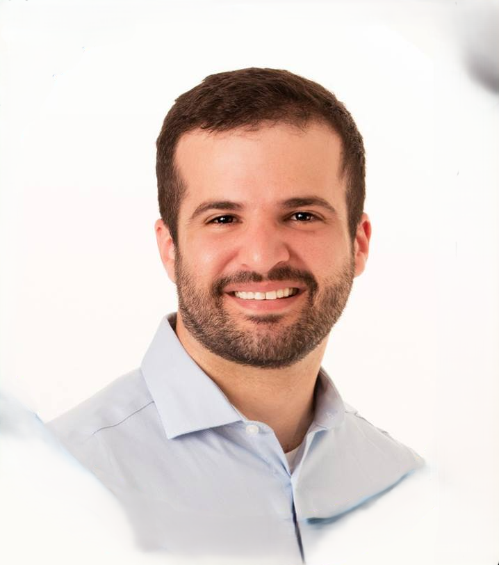 A smiling man with short brown hair and a beard, wearing a light blue dress shirt, posing against a plain white background.
