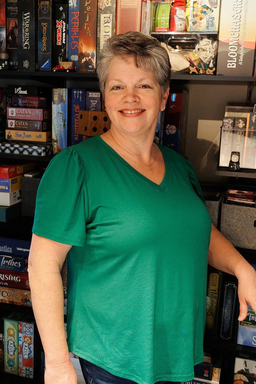 A woman with short gray hair smiling, wearing a green top, standing in front of a bookshelf filled with board games, books, and miscellaneous items.