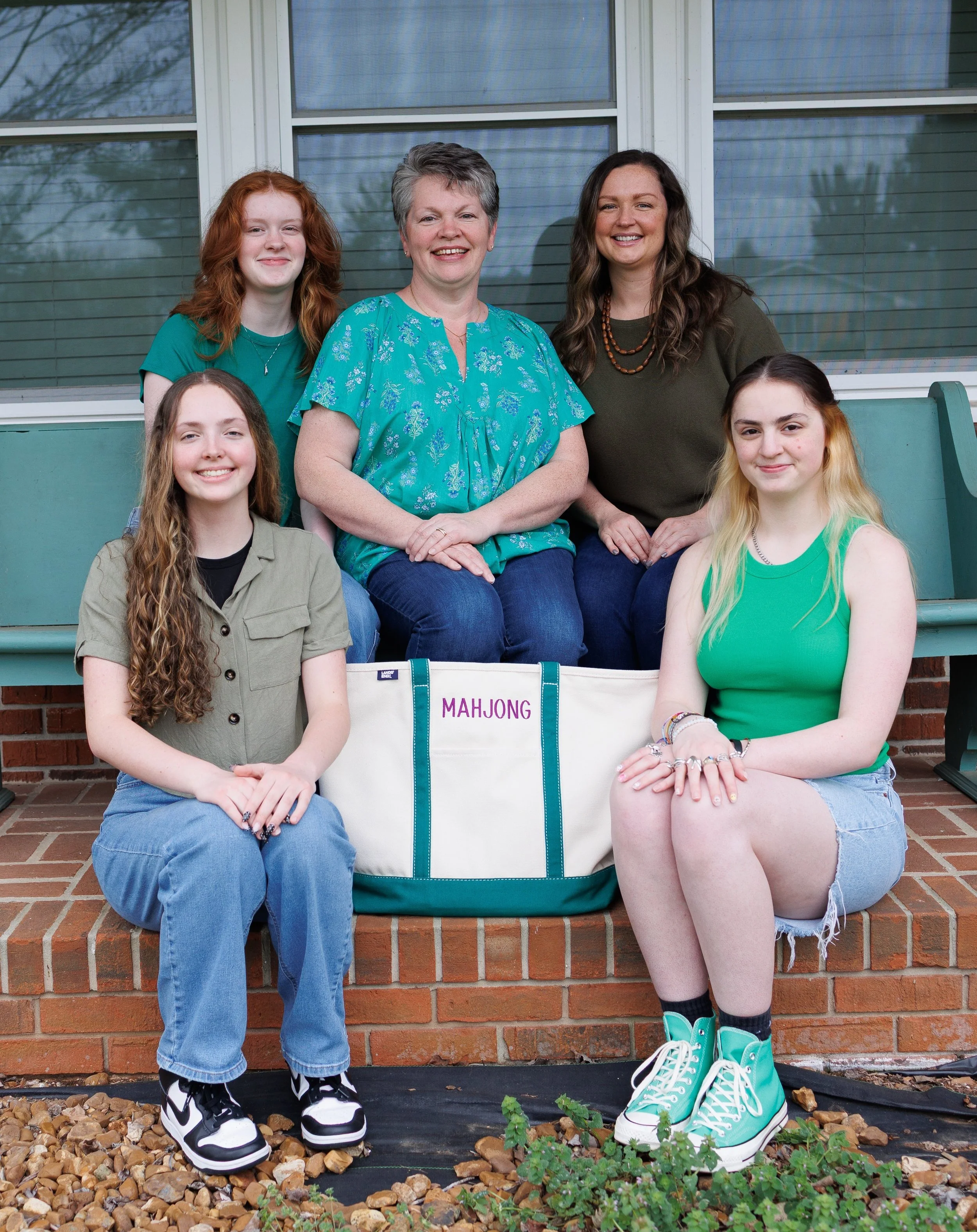 A group of six women and girls sitting and standing on a brick porch in front of windows, with a bag labeled 'MAHJONG' in front, smiling at the camera.