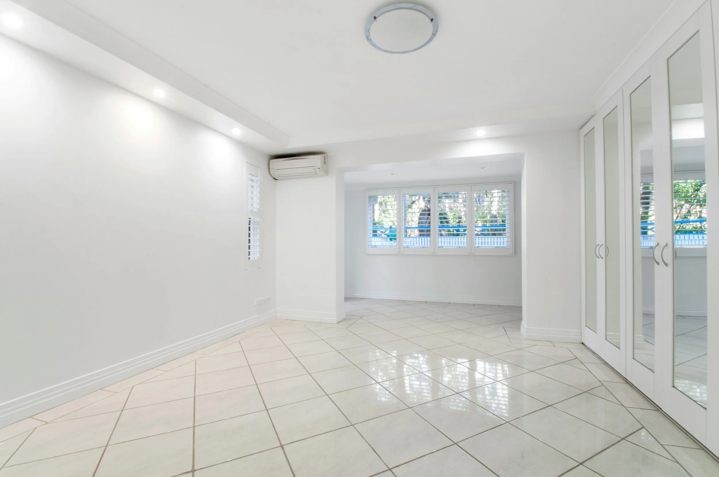Empty, bright living room with white walls, tiled floor, large windows with blinds, an air conditioner, and mirrored closet doors.