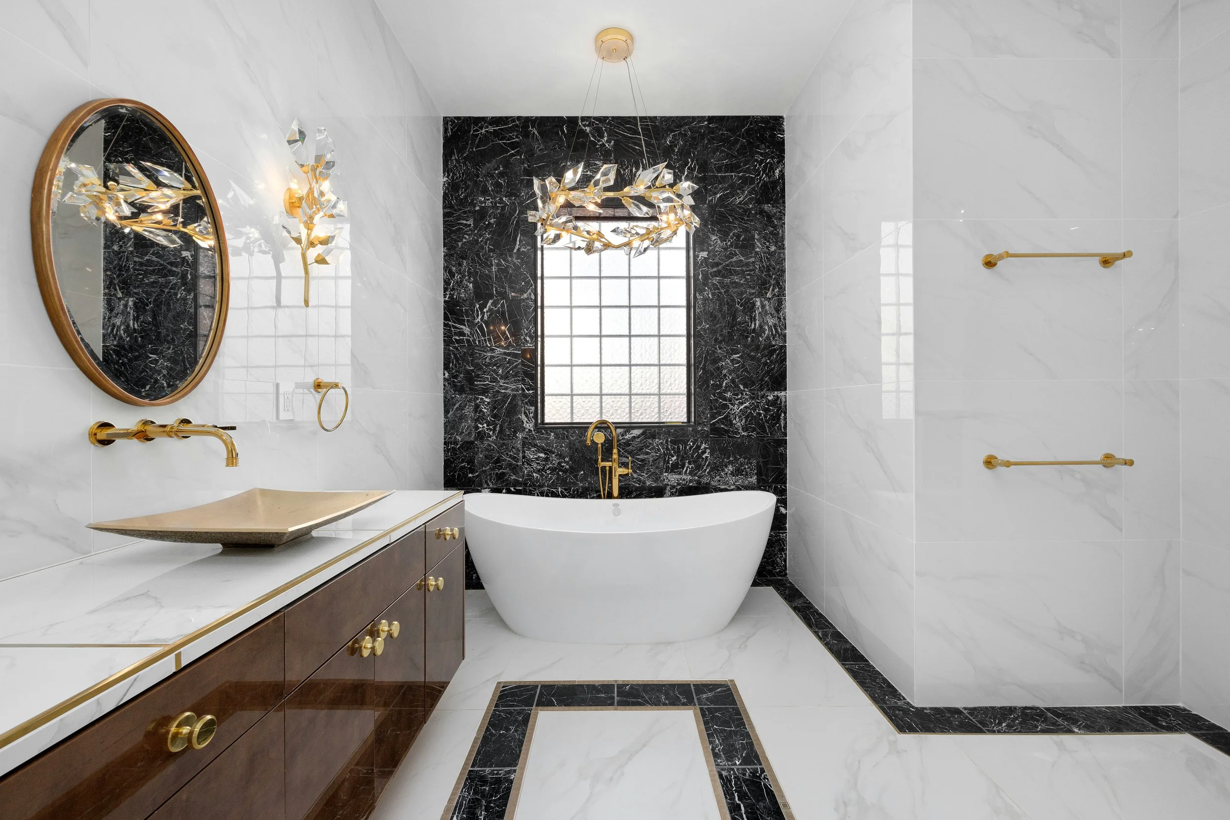 Modern bathroom with white marble walls and floor, black marble accent wall behind a white bathtub with gold fixtures, a dark wood vanity with a white marble countertop, a gold-framed mirror, decorative gold wall sconce, and gold towel bars.