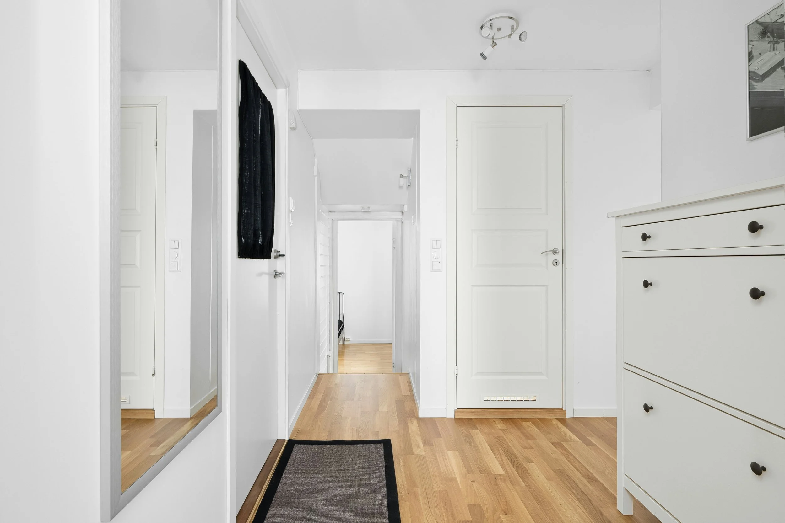 A bright, minimalist hallway with white walls and light wooden flooring, featuring a large wall mirror, a black curtain on a door, a white dresser with black knobs, and a framed photo on the wall.