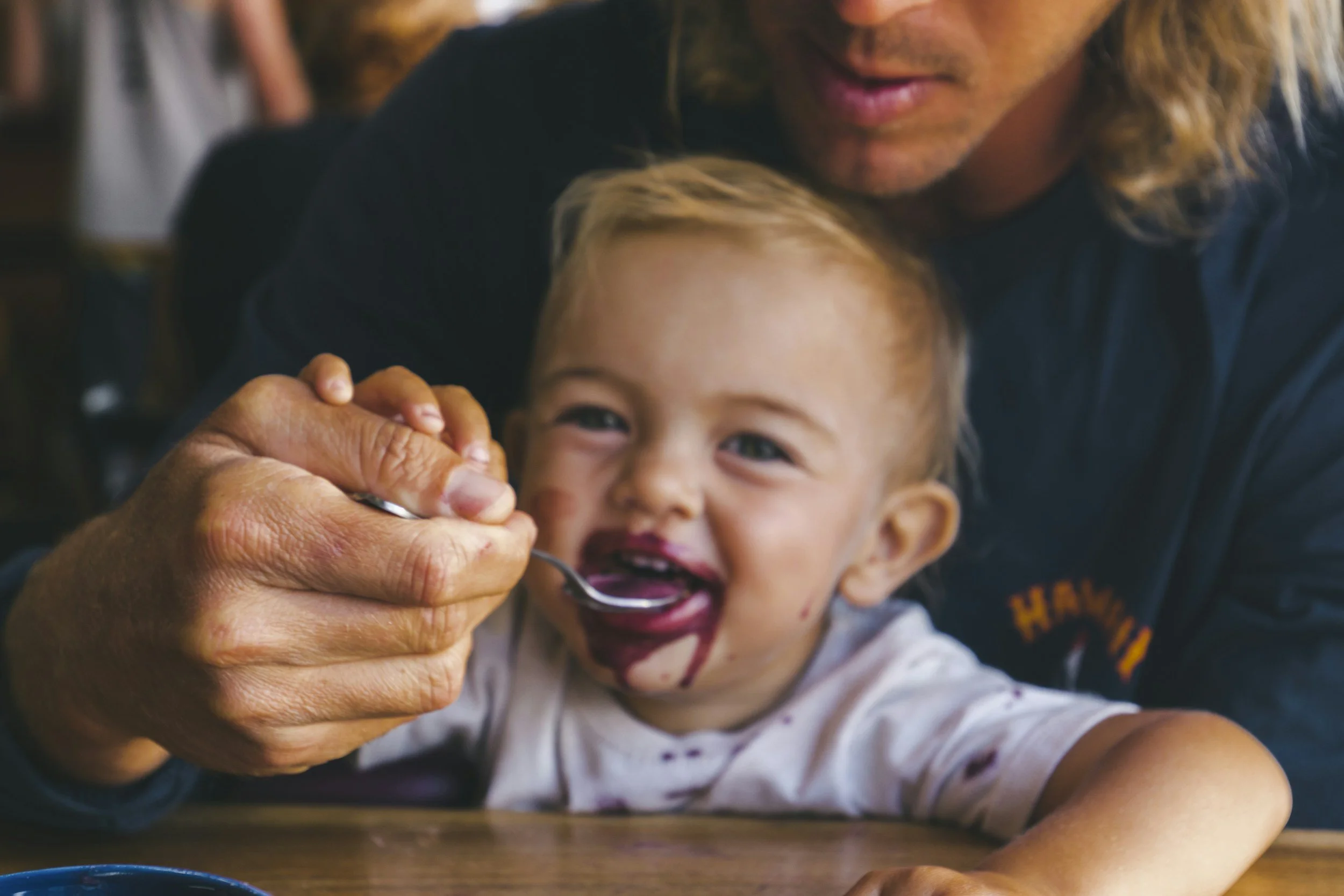 A young child with messy face and lips, enjoying food with the assistance of an adult, likely a family member, in a warm indoor setting.