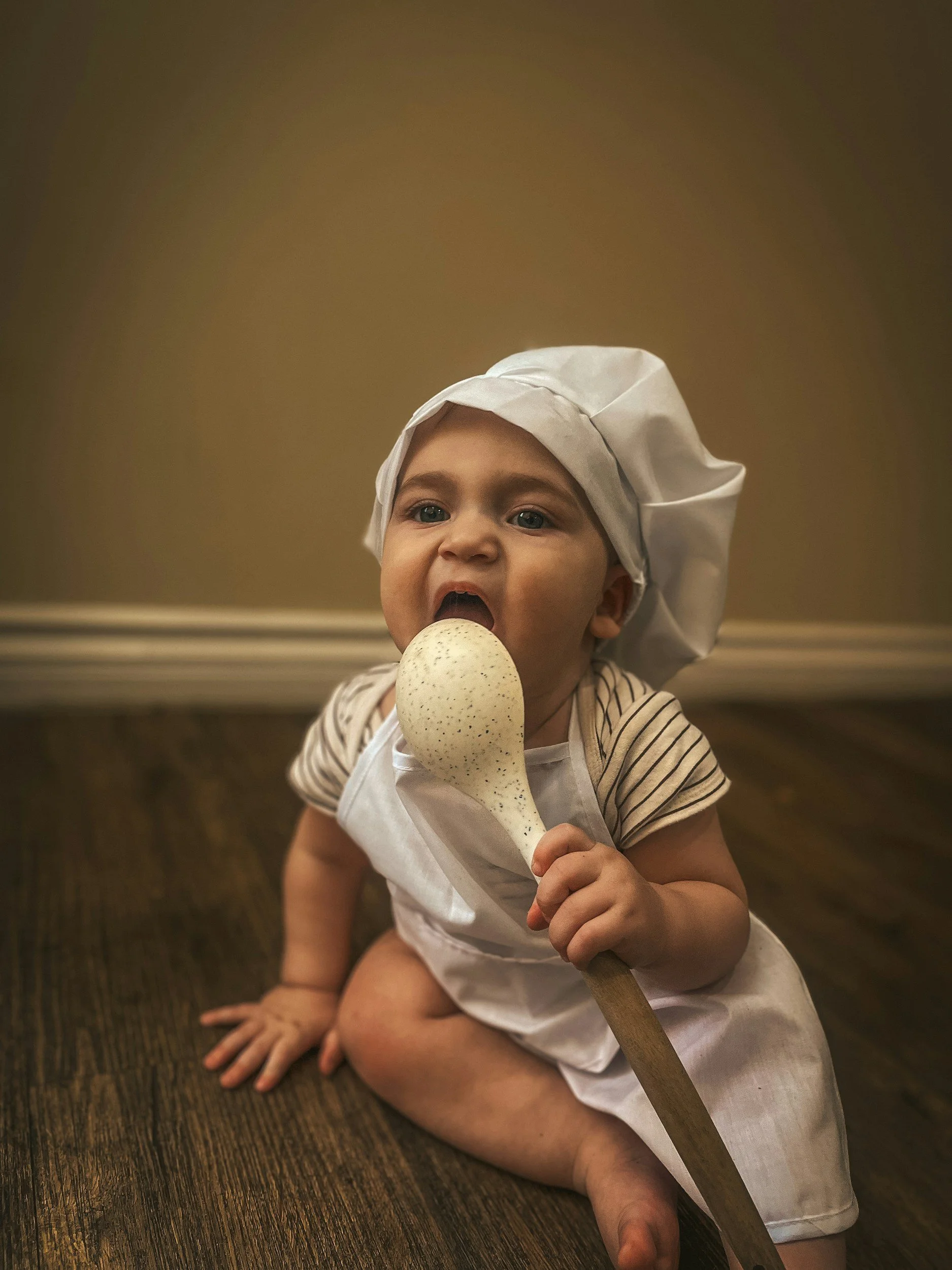 A young child dressed as a baker, wearing a white chef's hat and apron, is sitting on the wooden floor and pretending to eat a large spatula.