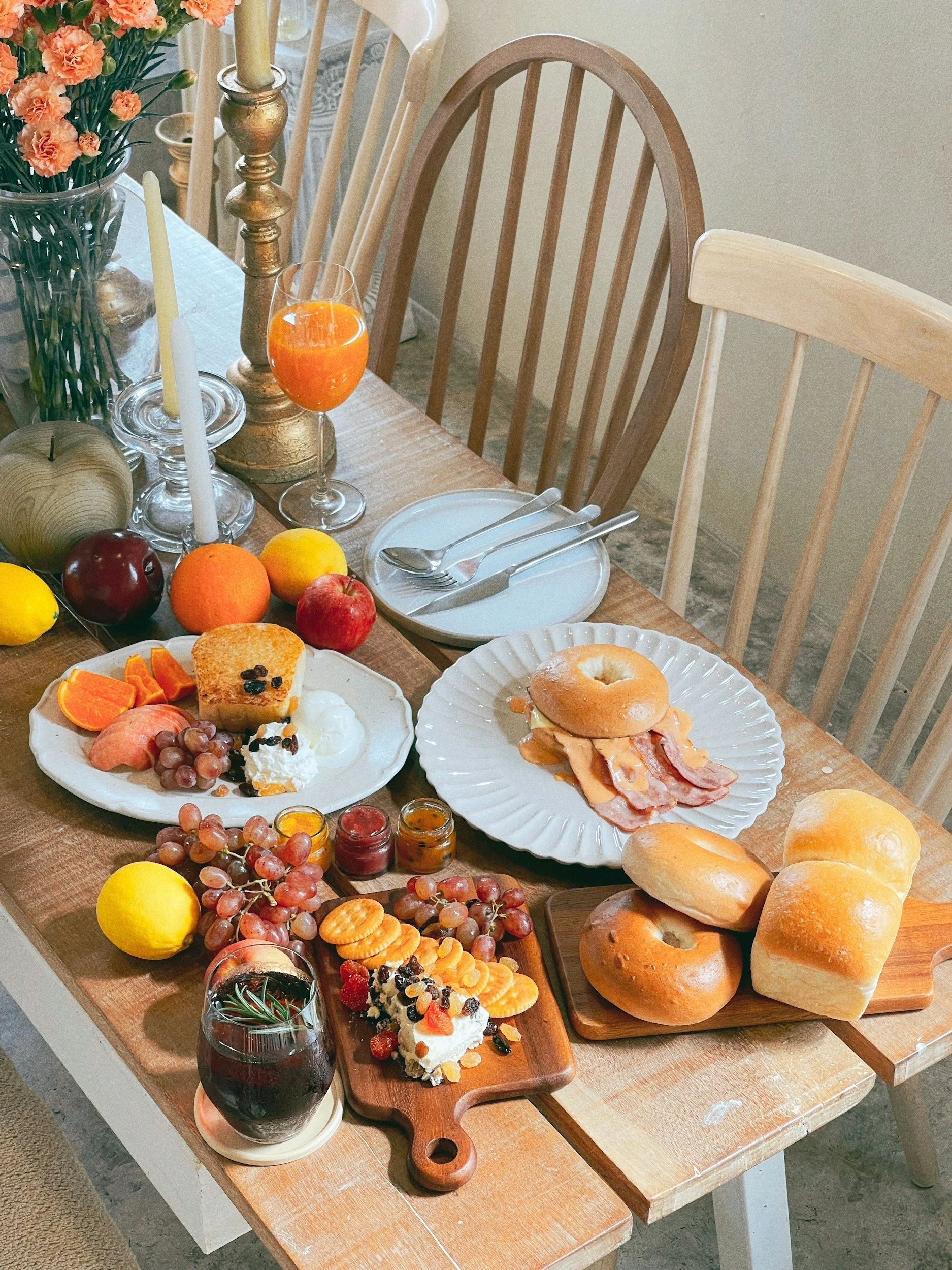 A breakfast table with assorted foods including bagels, bread rolls, bacon, eggs, fruit (grapes, apples, oranges, lemon), cheese, cereal, jam, and a glass of orange juice. There are also candles, a flower vase, and tableware such as plates, cutlery, and glasses.