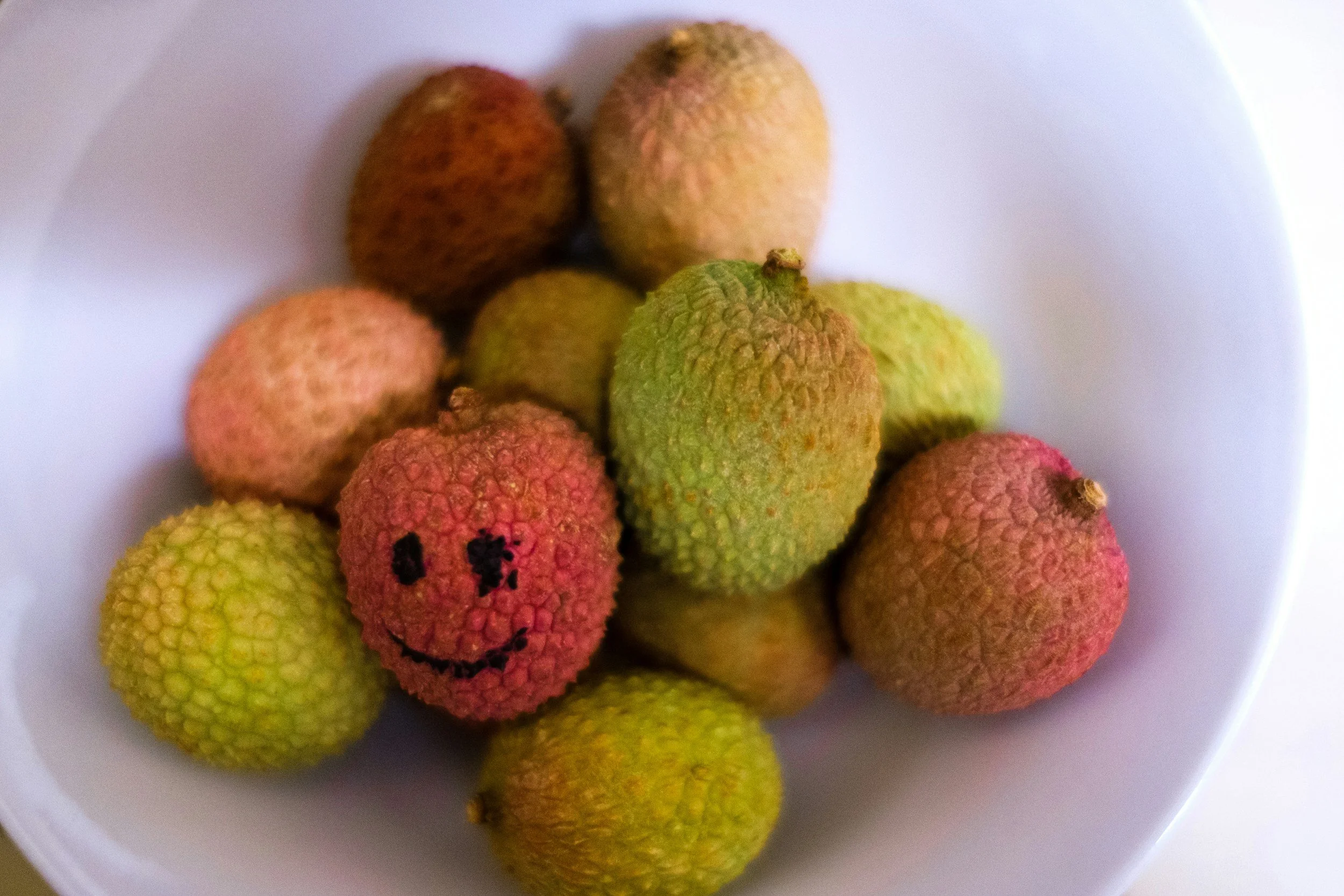 A white bowl containing several lychee fruits, with one lychee having a smiling face and eyes drawn on it, and a happy mouth, giving it a cartoonish appearance.