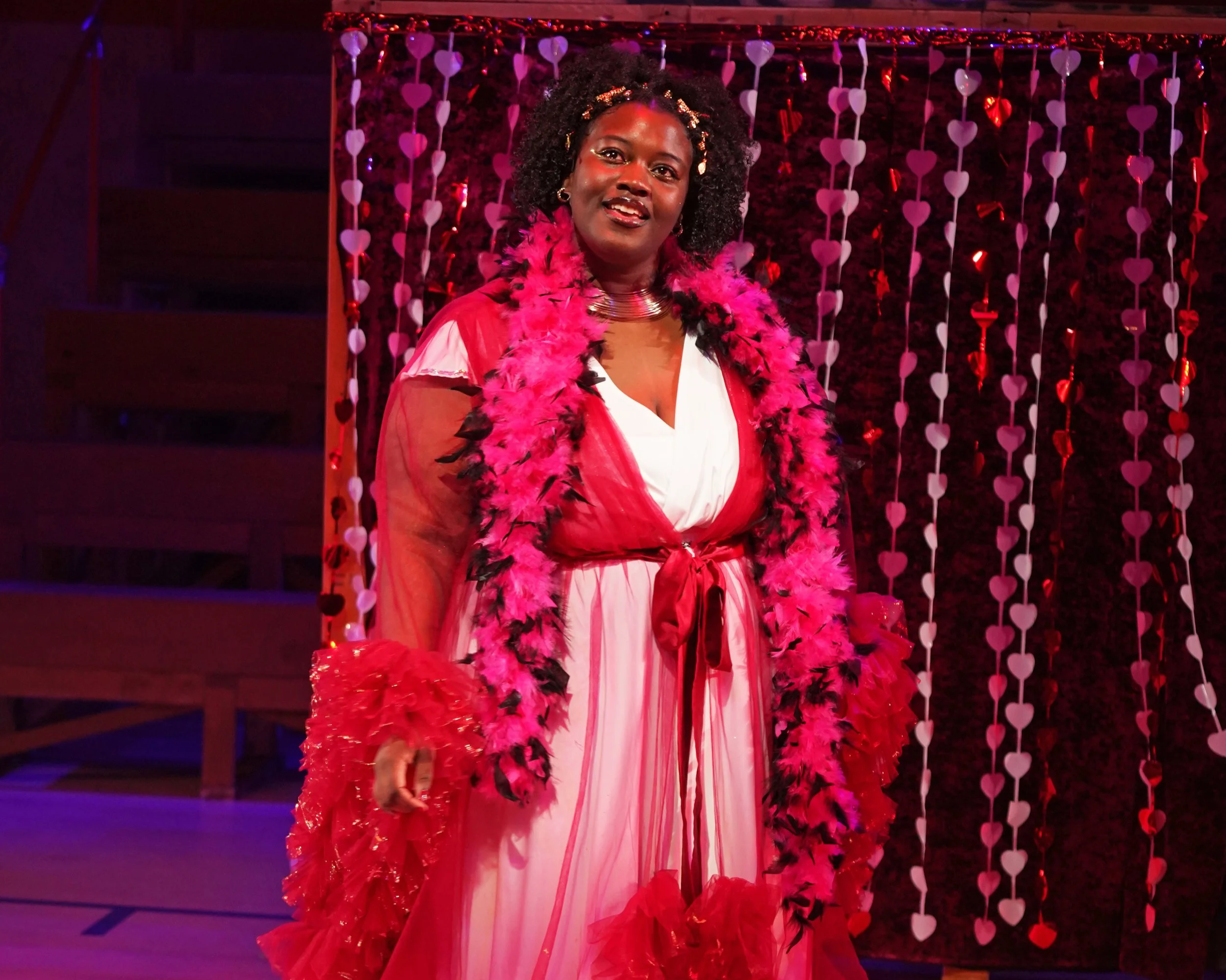 A woman dressed in a pink and white dress with a pink feather boa and red gloves, standing in front of a pink heart-themed decorated backdrop.