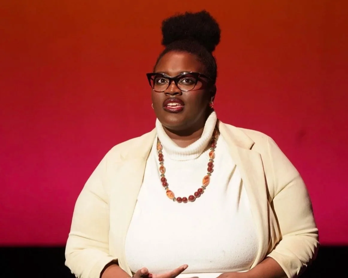 A woman with glasses, wearing a white turtleneck and a cream blazer with a colorful beaded necklace, standing in front of a red background.