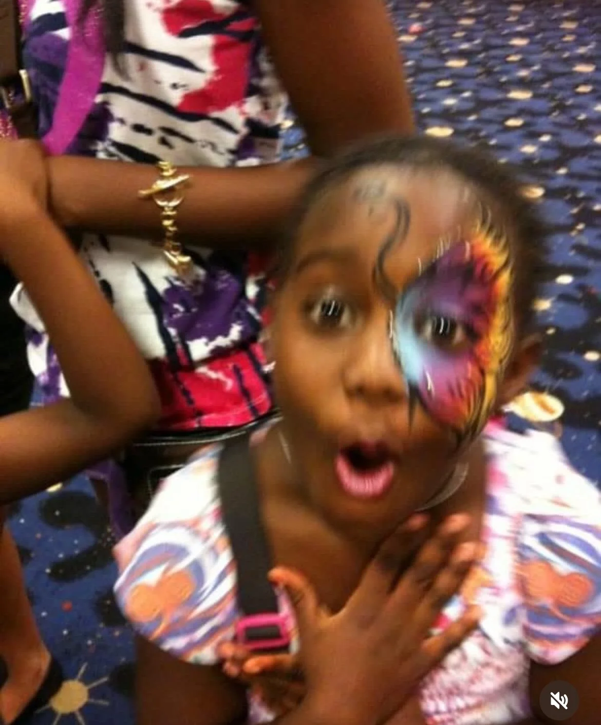 A young girl with a butterfly face painting, showing an amazed expression, clutching her chest with hands crossed, on a colorful patterned carpet.