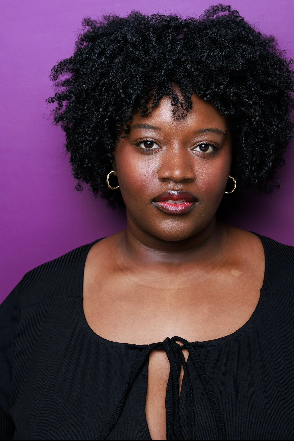 Close-up portrait of a young Black woman with curly black hair, wearing gold hoop earrings and a black top with a front tie, standing against a purple background.