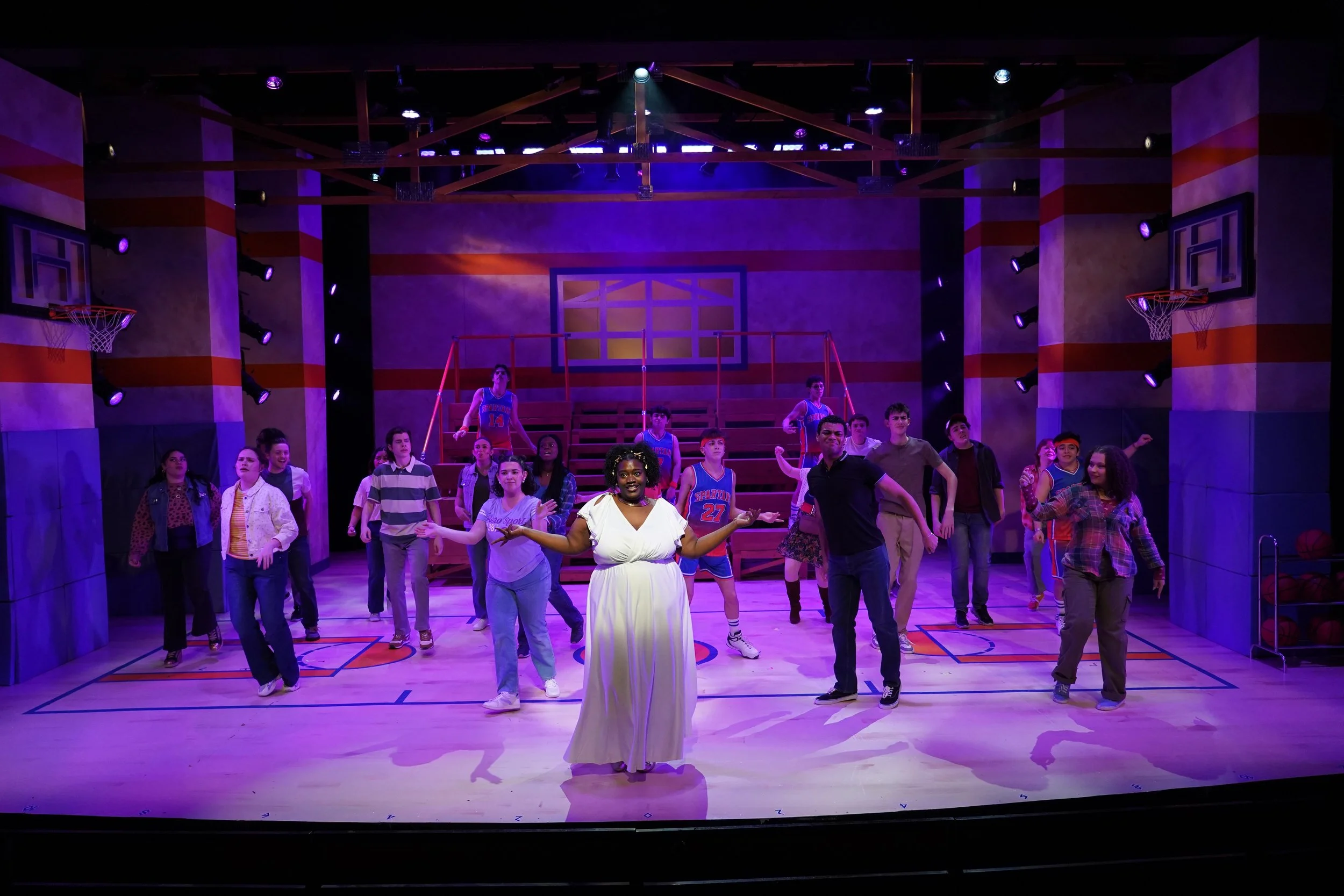 Stage scene of a musical or play set in a gymnasium with a basketball court, with performers in casual clothing, some in sports jerseys, on stage with purple lighting.