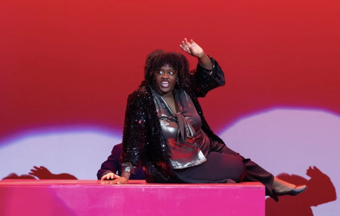 A woman performing on stage, sitting on a pink platform with a red background, wearing a shiny black top and a sequined jacket.