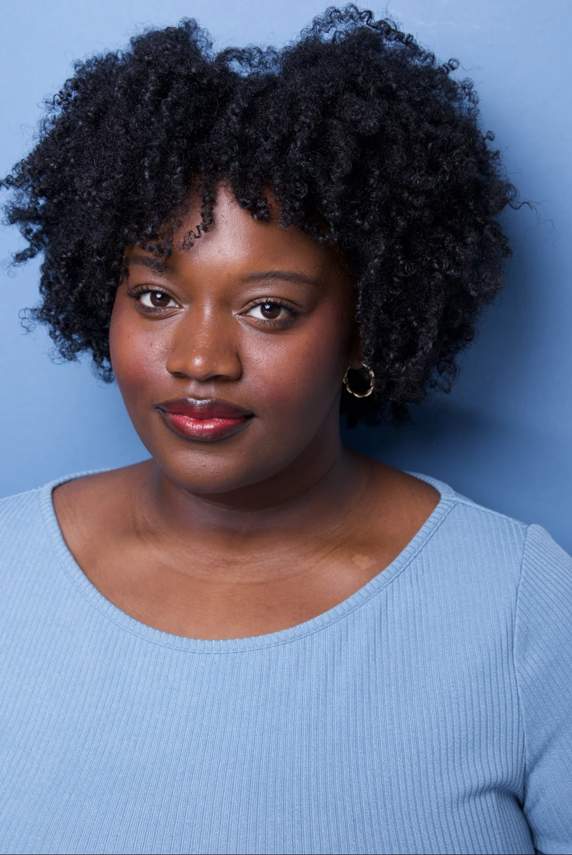 Portrait of a woman with dark curly hair, wearing a light blue top and gold hoop earrings, standing against a blue background.