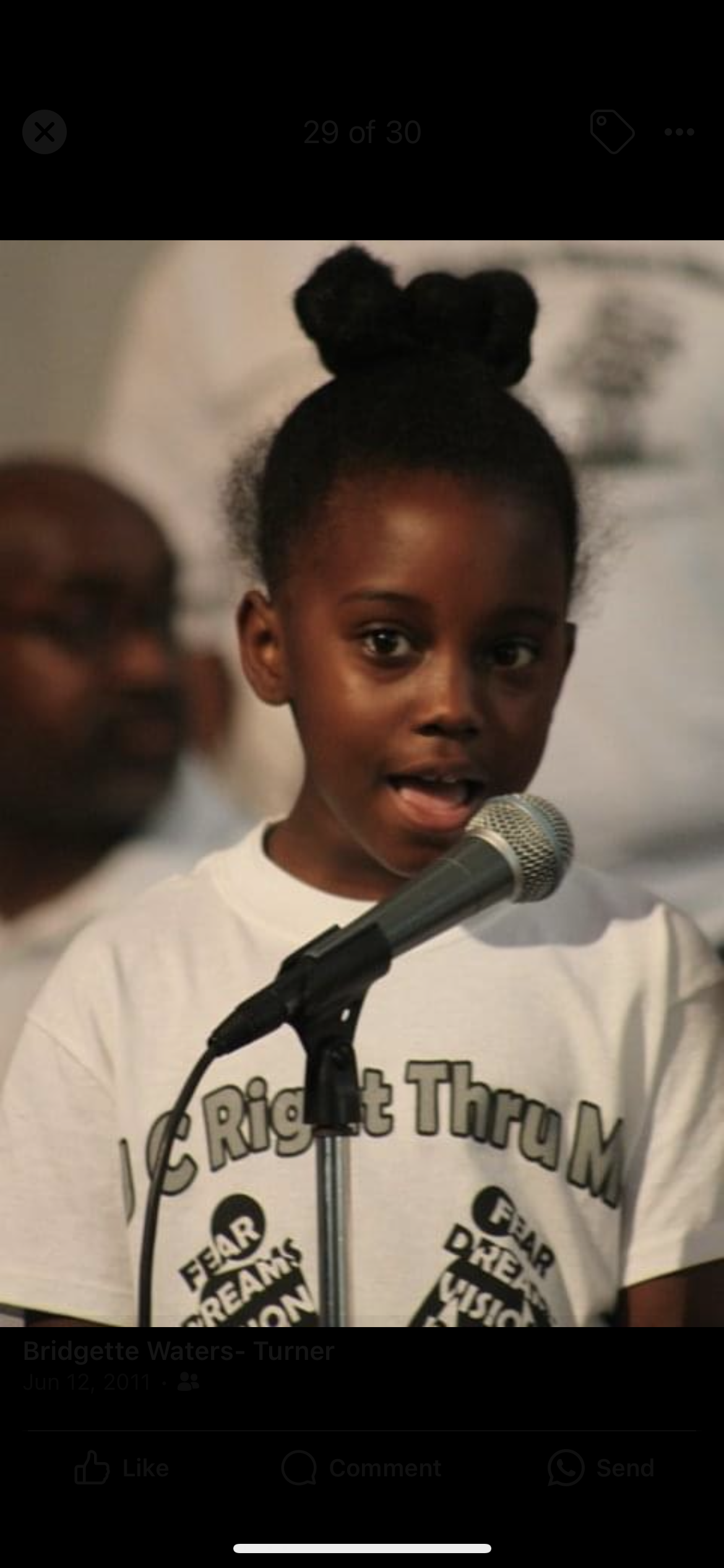 A young girl with dark hair styled in two buns, speaking into a microphone at a public event, wearing a white T-shirt that has a graphic with words about dreams and visions.