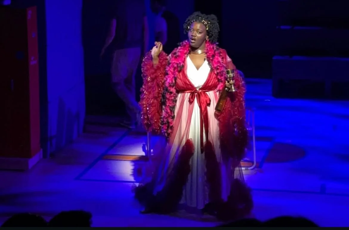 A woman on stage wearing a white and red dress with a red tie and a dramatic red feathered cape. She has curly hair and is holding a golden object in her left arm. The stage lighting is blue and purple.