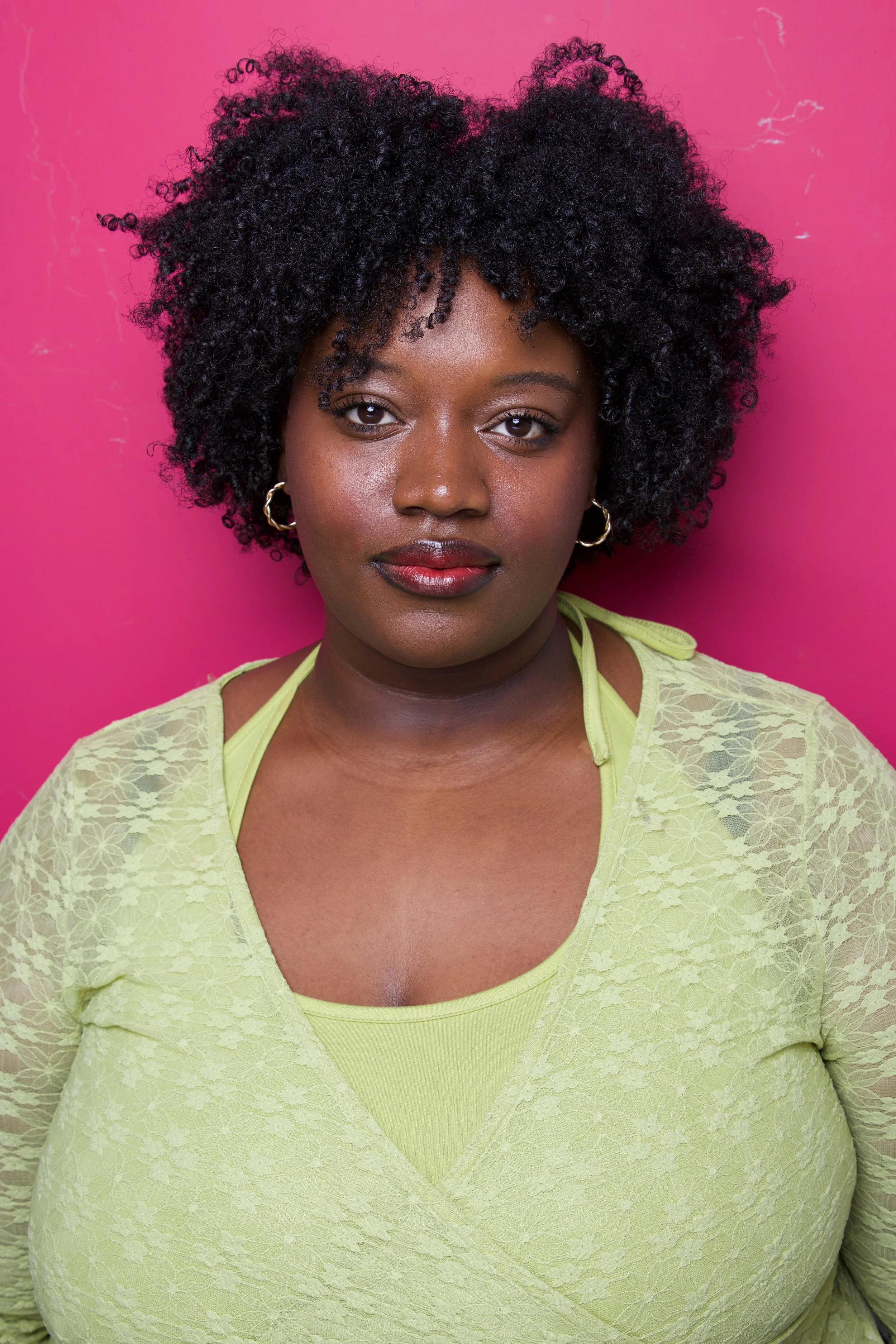 Portrait of a woman with dark, curly hair, wearing hoop earrings and a yellow lace top, in front of a pink background.