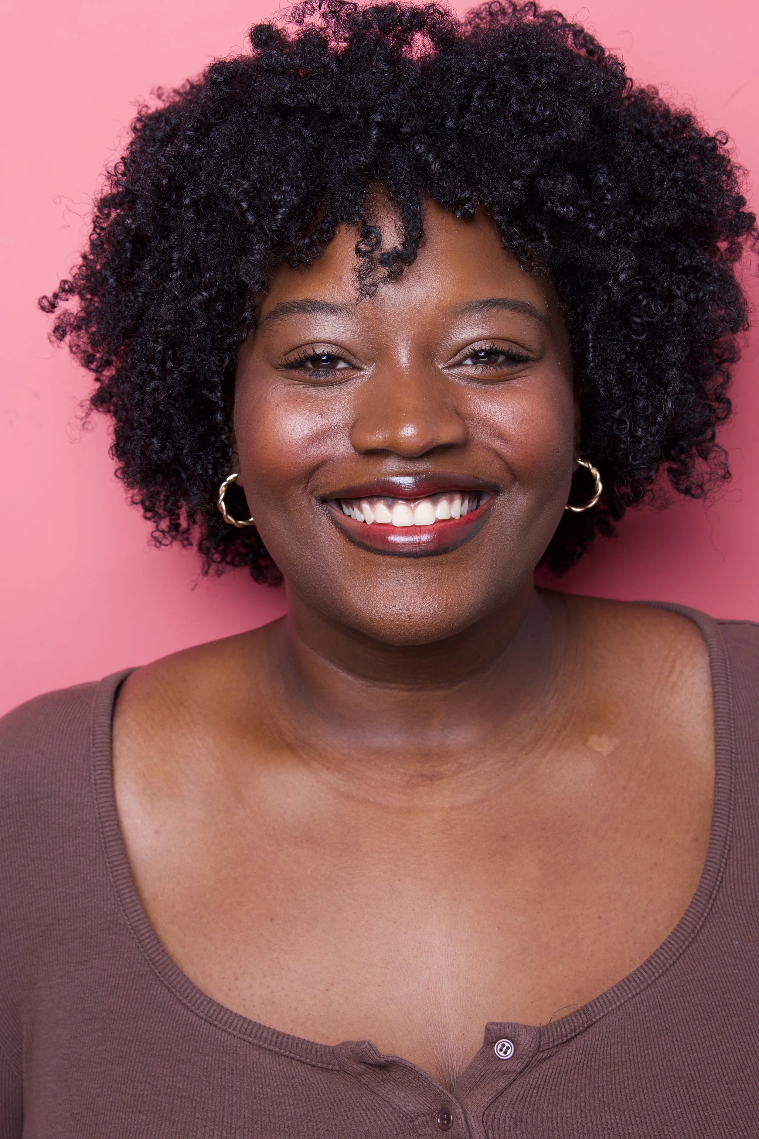 Close-up portrait of a smiling Black woman with short curly hair, wearing gold hoop earrings, a brown shirt, against a pink background.