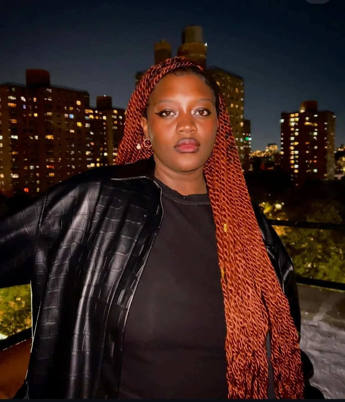 A woman with red braided hair standing outdoors at night in front of city buildings with lights shining from the windows.