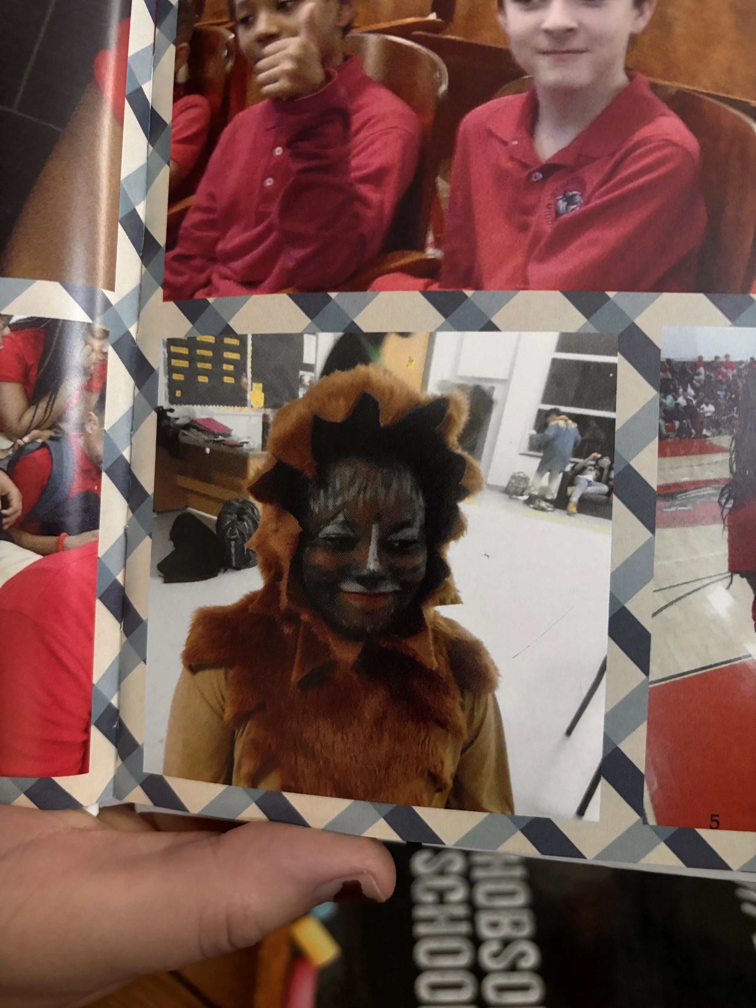 A person holding a scrapbook open to a page with two photos. The top photo shows two boys in red shirts sitting on wooden chairs, one giving a thumbs-up. The bottom photo features a person in a lion costume with face paint resembling a lion, smiling 