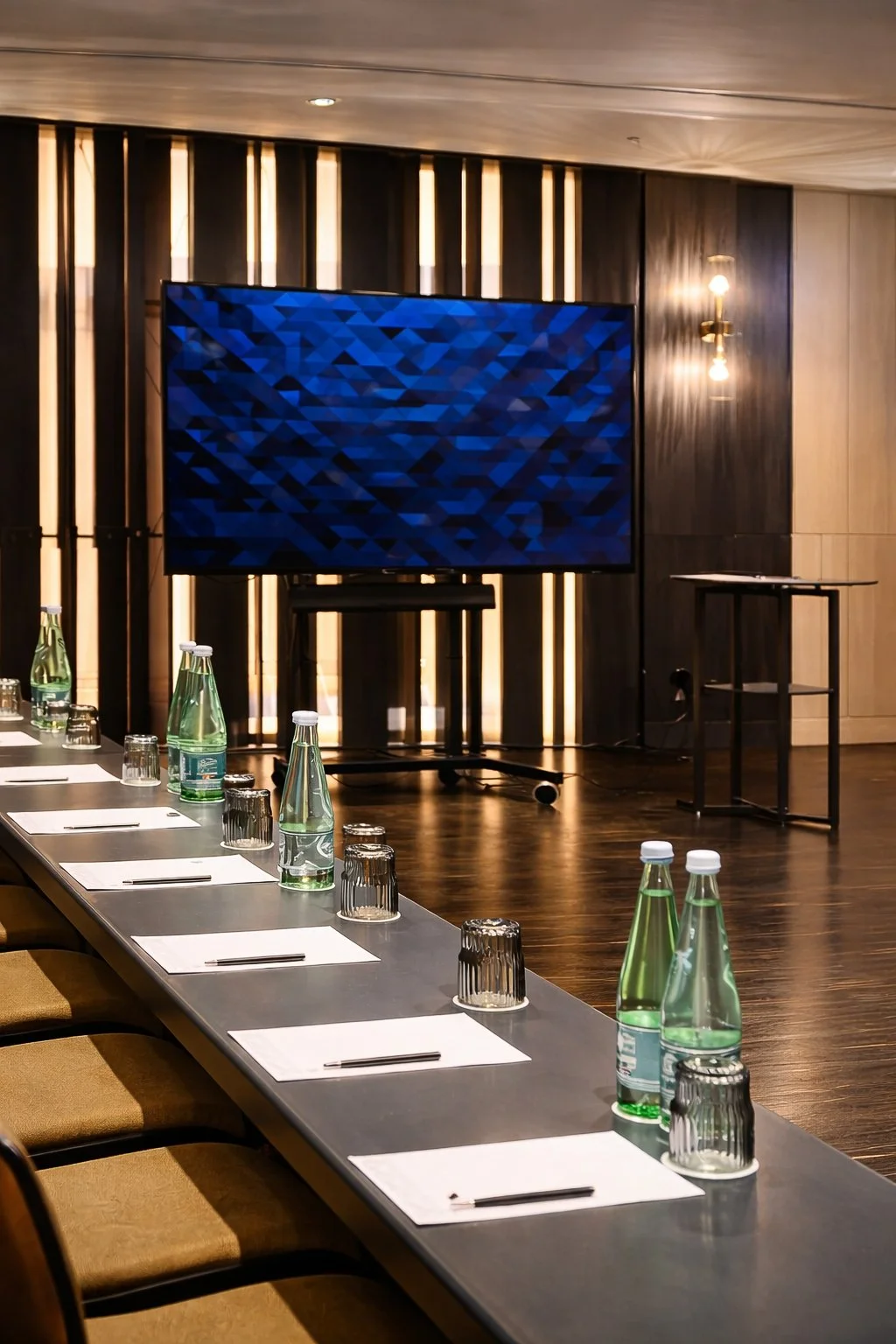 Conference room setup with a long table, water bottles, glasses, notepads, and a large screen at the front.