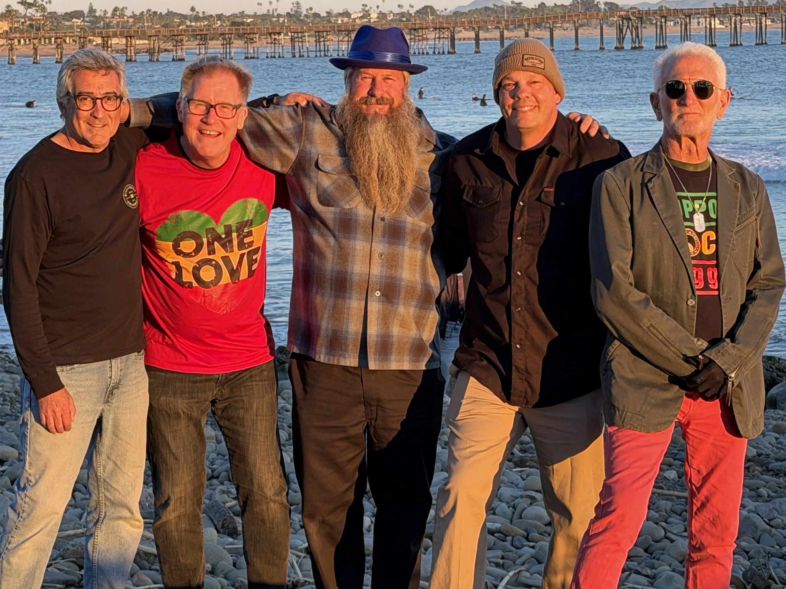 Six men standing together on a rocky beach with a pier and ocean in the background, during sunset.