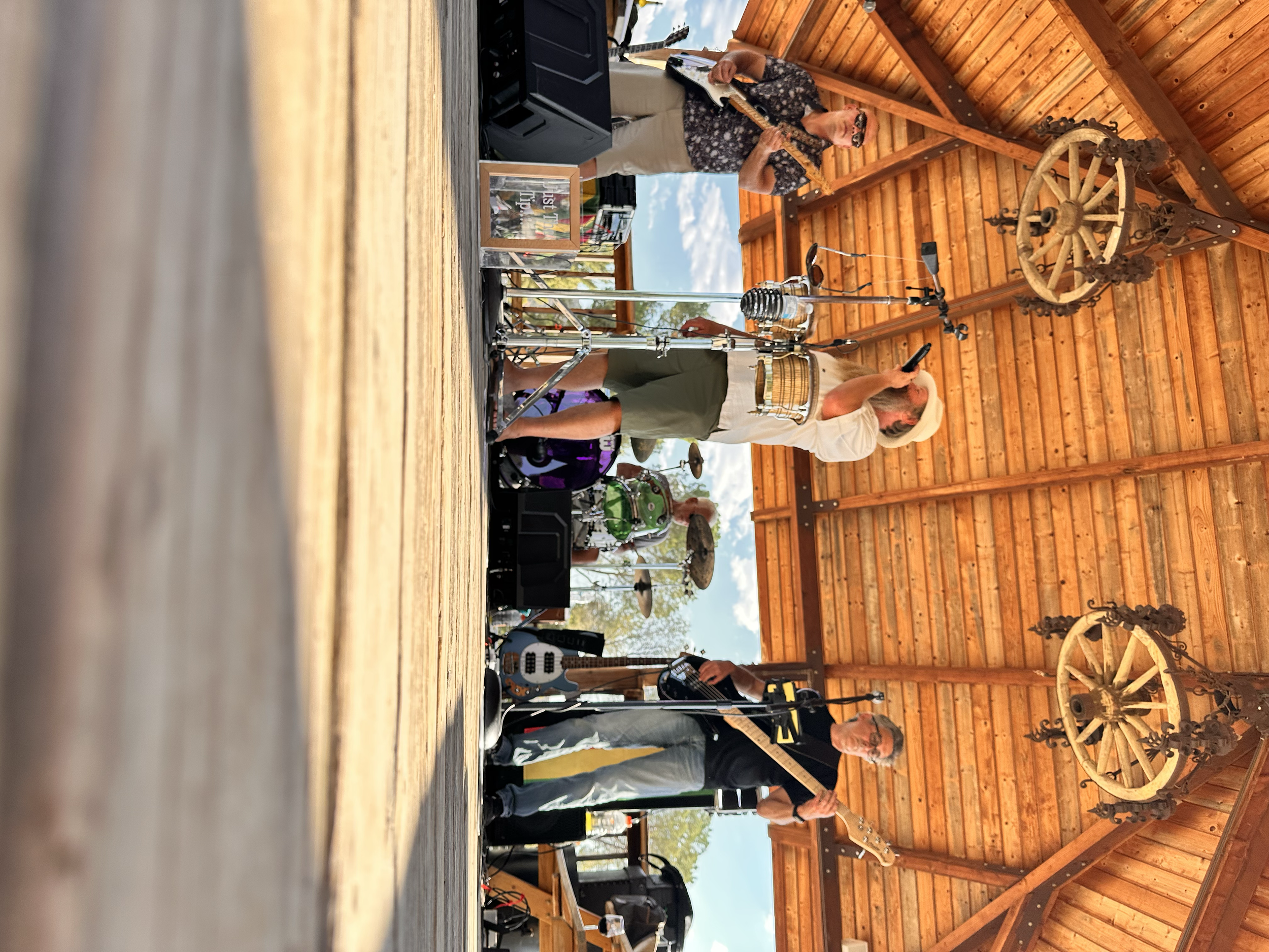 Two musicians playing guitar and drum set on a wooden stage with a wooden backdrop, under a peaked roof, during daytime.