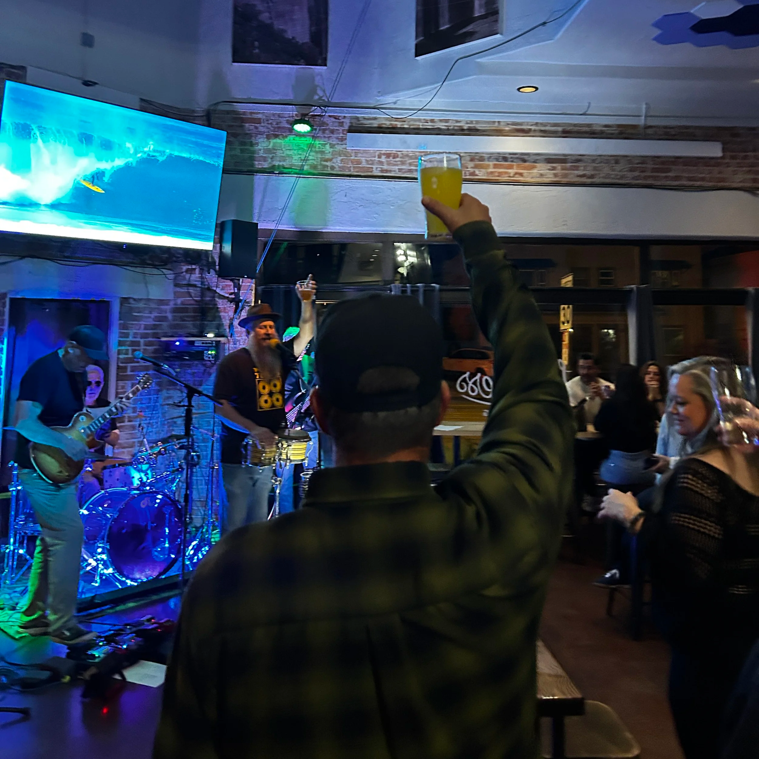 People at a bar watching a live band perform, with one person in the foreground raising a yellow drink in a glass.
