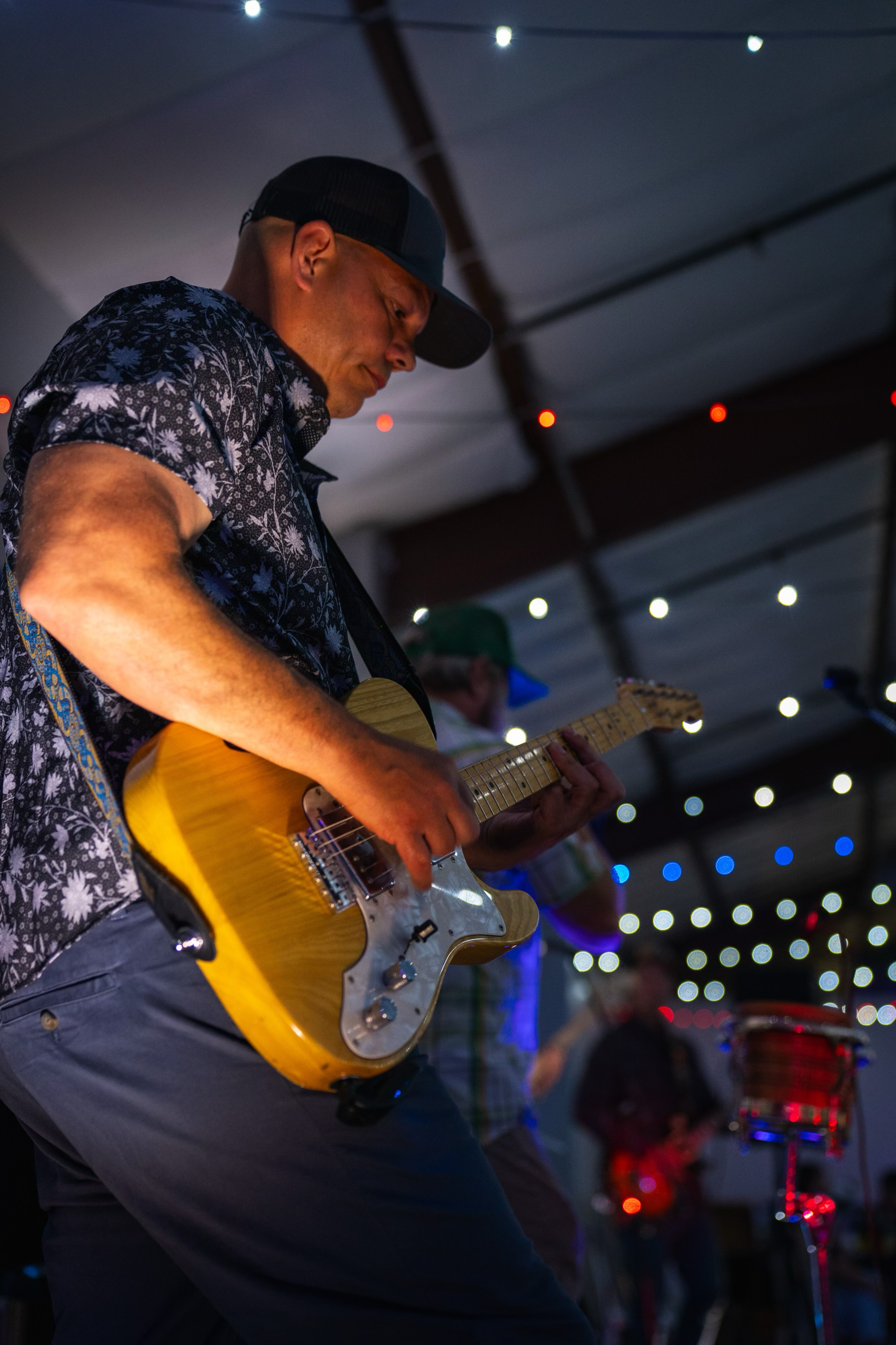 A man playing an electric guitar on stage under string lights, wearing a black cap and a printed shirt.