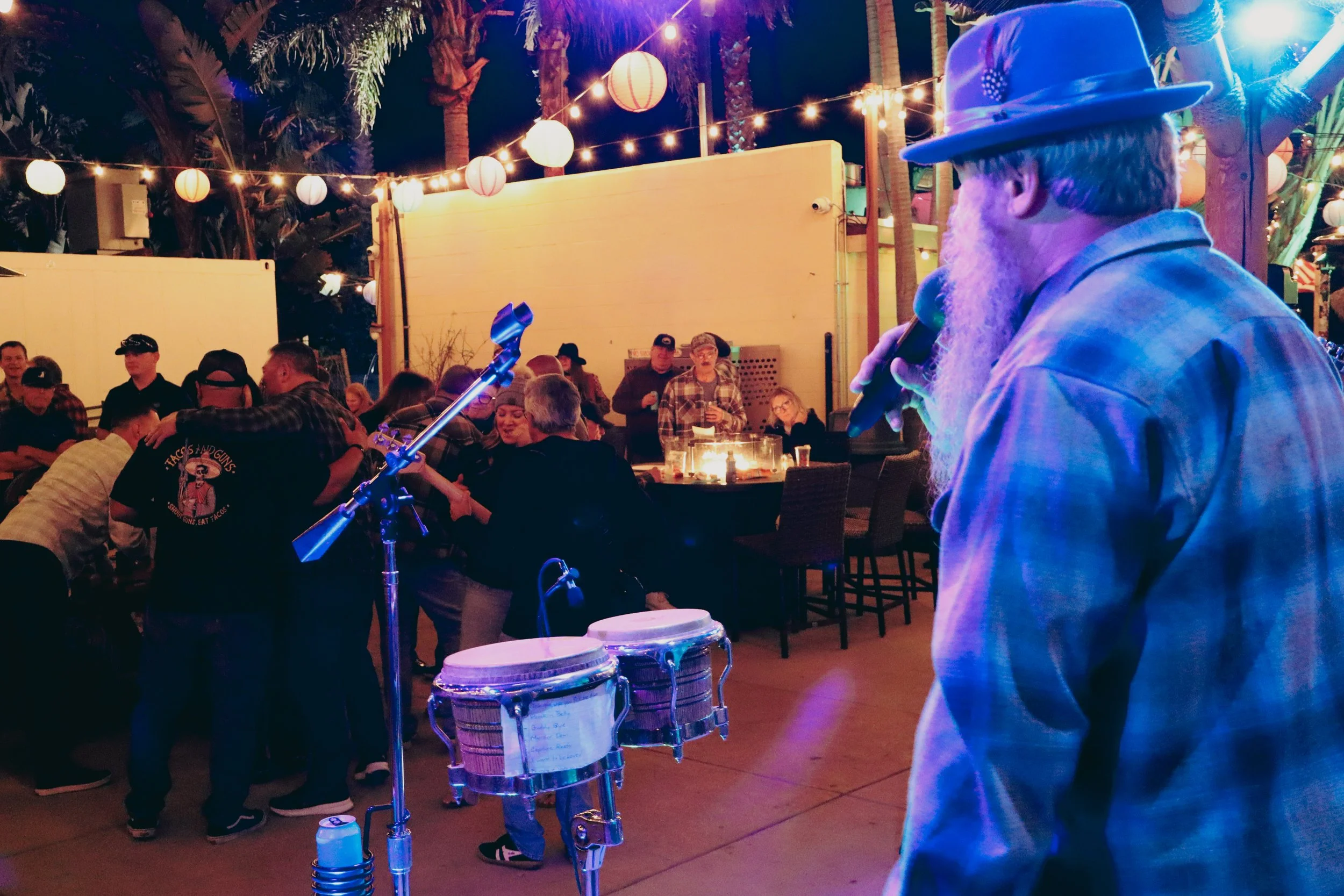 A man with a white beard wearing a hat and plaid shirt holding a microphone and singing at an outdoor party, with people dancing and socializing under colorful string lights and paper lanterns.