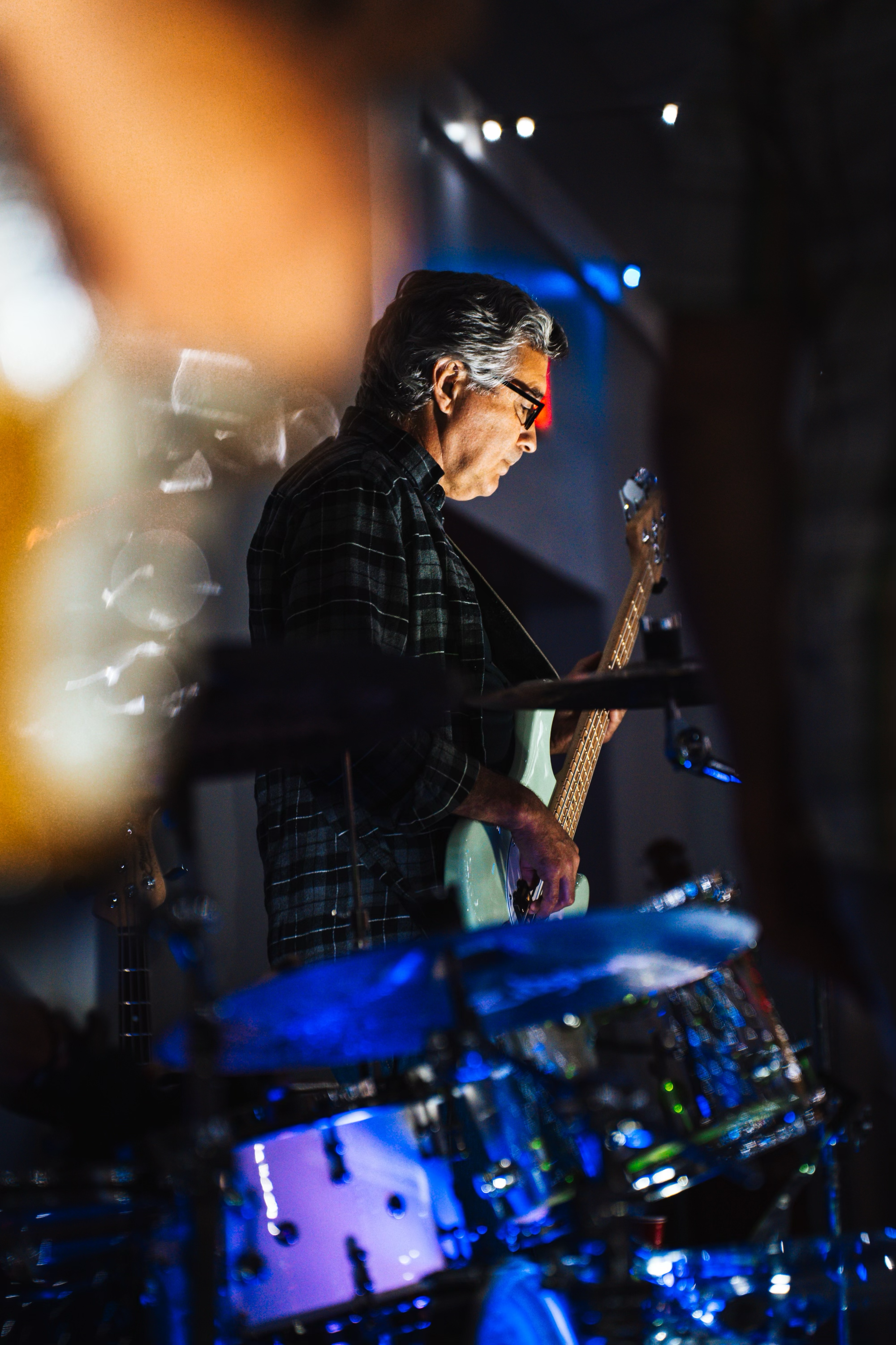 A man playing an electric guitar next to a drum set, with colorful stage lighting.