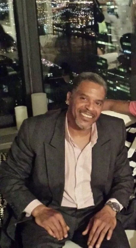 A man in a suit smiling at an indoor restaurant with a cityscape view at night in the background.