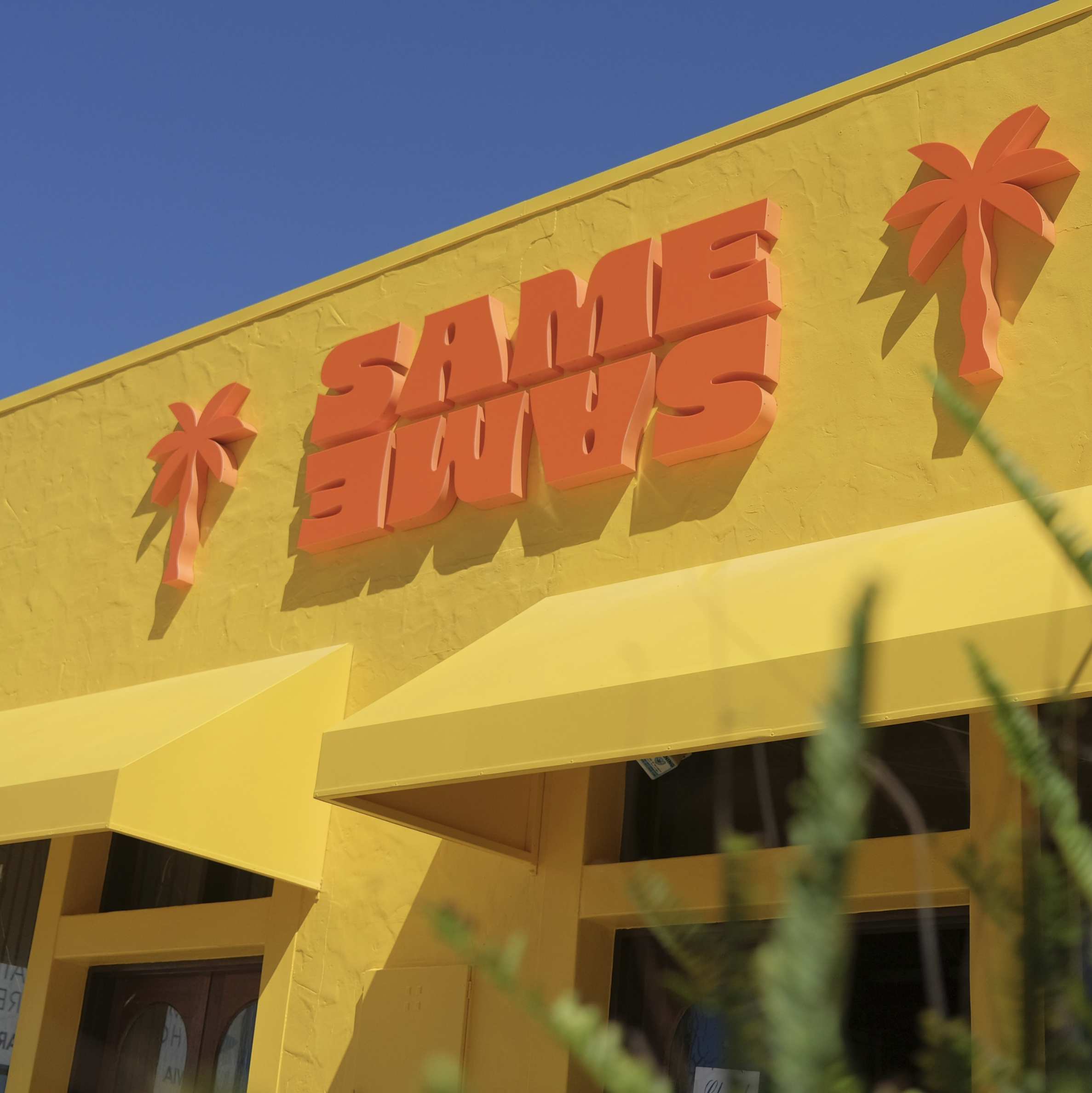 Same Same storefront signage featuring bold 3D fabricated letters in orange, mounted on a vibrant yellow facade with architectural awnings and high-impact retail branding.
