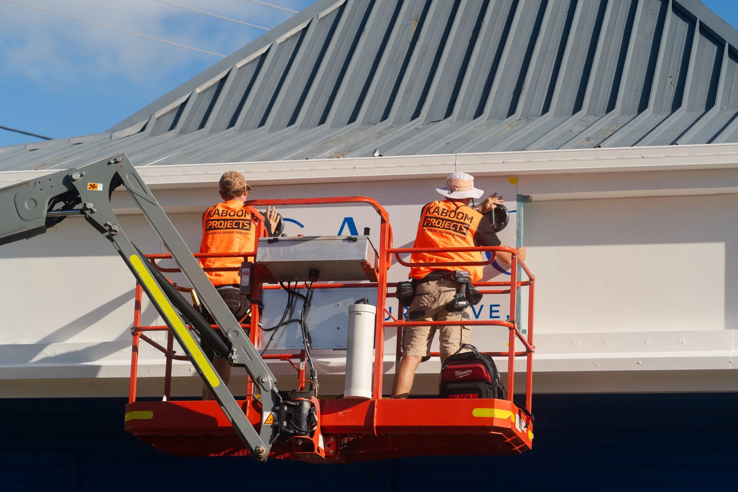 Kaboom Projects team installing external shopfront signage using an elevated work platform on a commercial building.